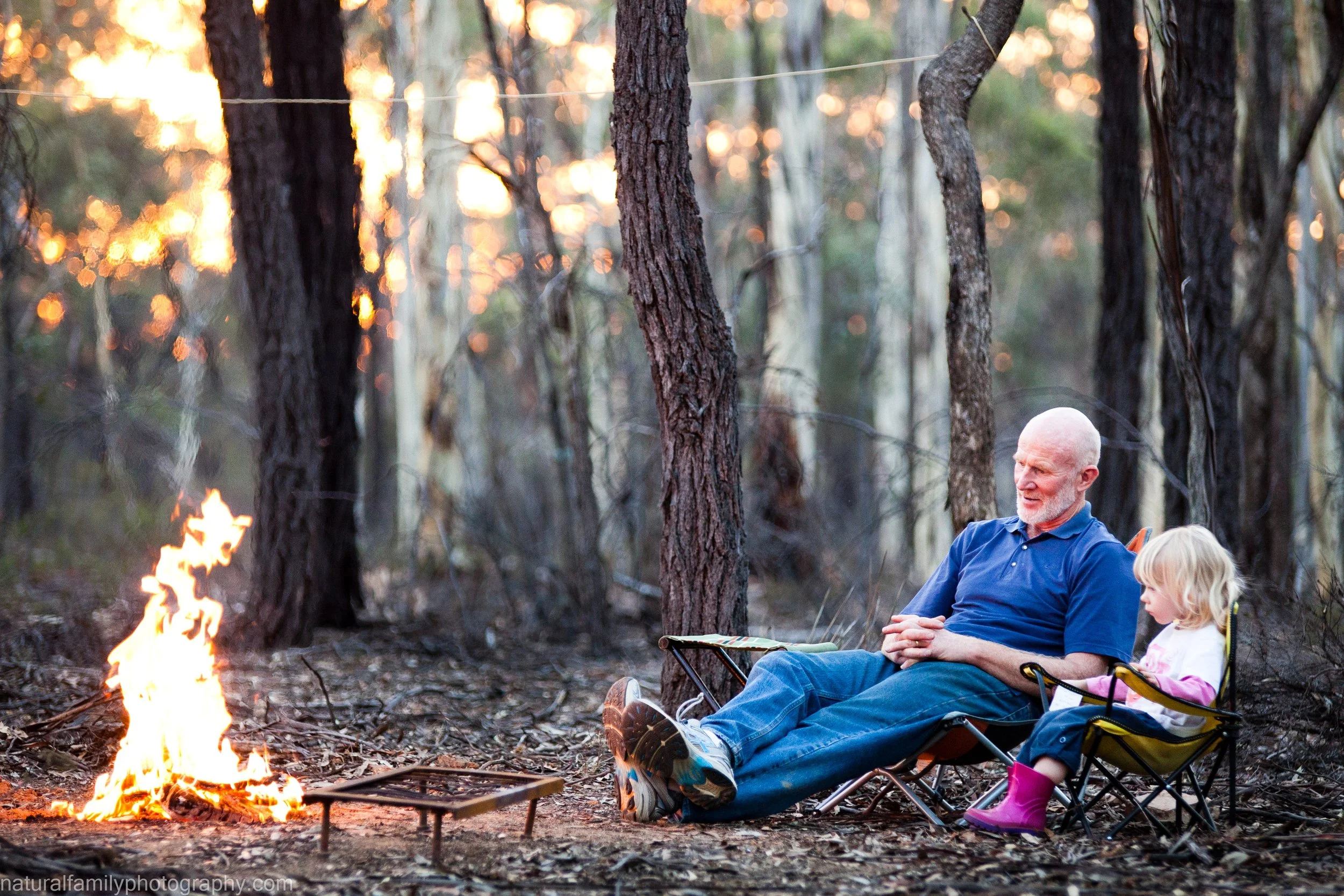 A grandfather and his granddaughter sit in camping chairs by a campfire in the Australian bush with tall trees at sunset. Location portrait by Natural Family Photography, Melbourne.