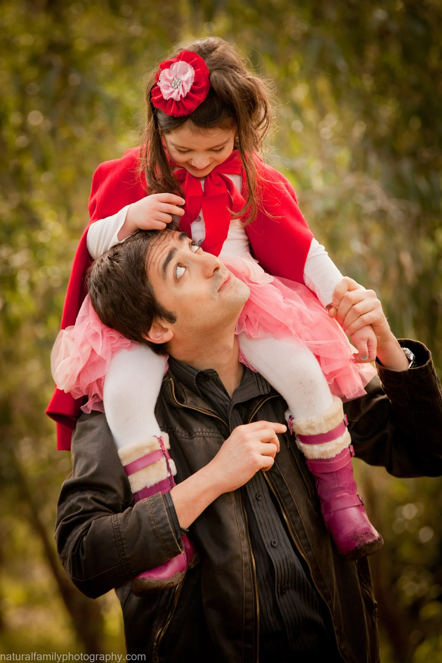 A man carrying a young girl on his shoulders while walking outdoors, surrounded by trees with autumn foliage.