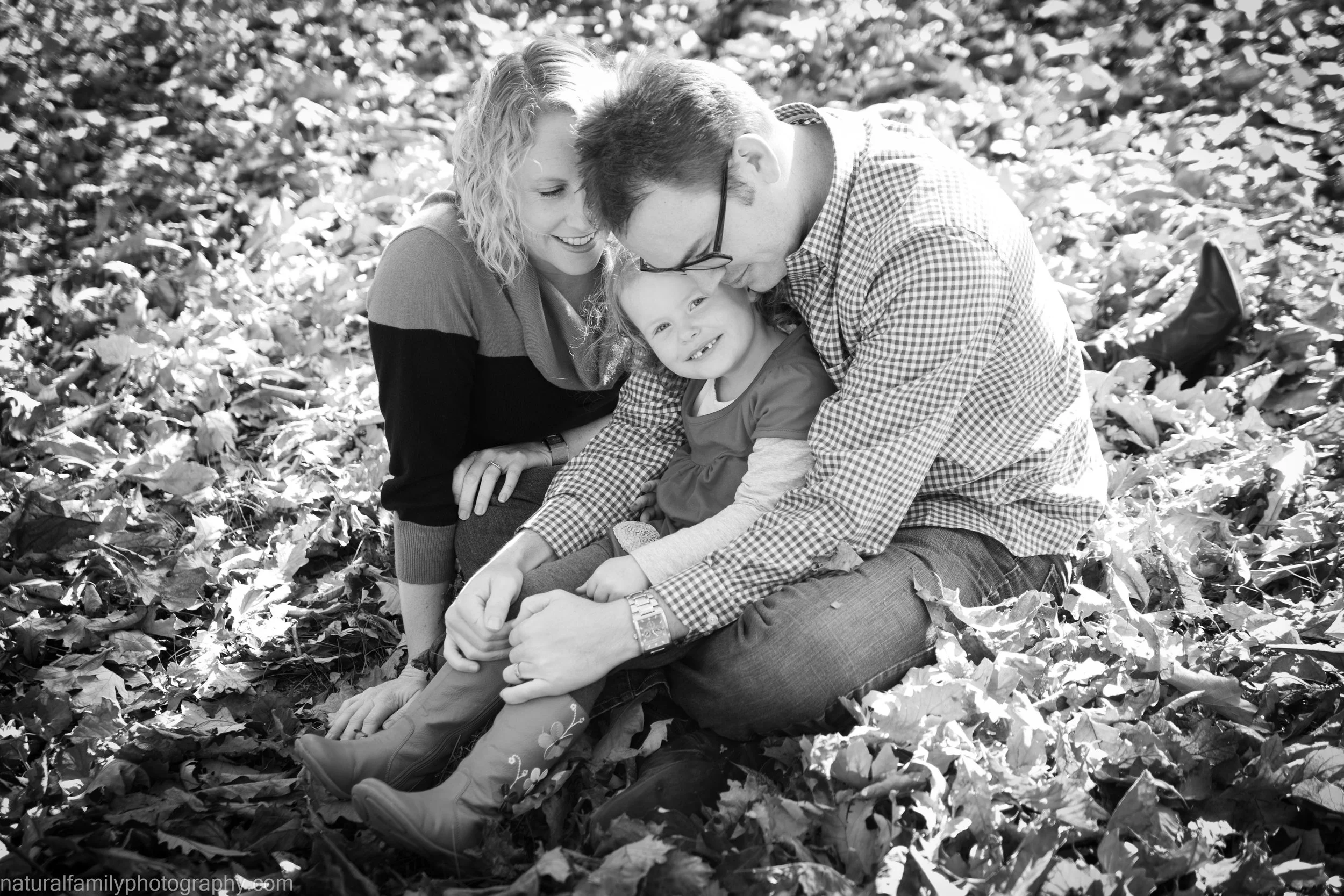A family sitting on a blanket of fallen leaves outdoors, with a woman, man, and young girl smiling and cuddling, sharing a happy moment.