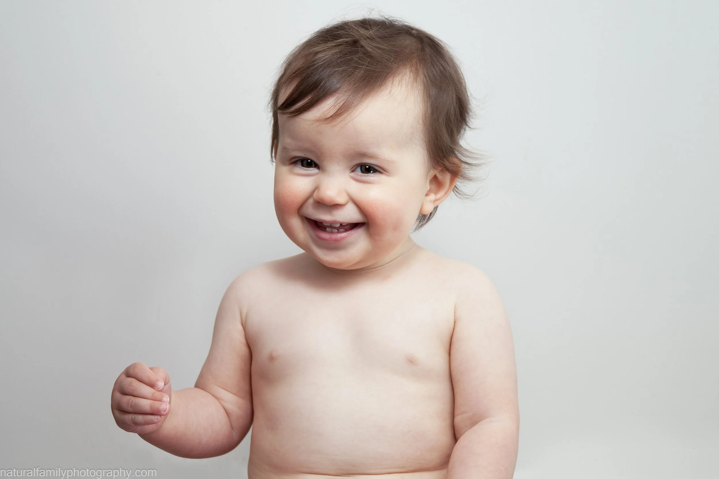 A smiling toddler with short brown hair and light skin, shirtless, standing against a plain white background.