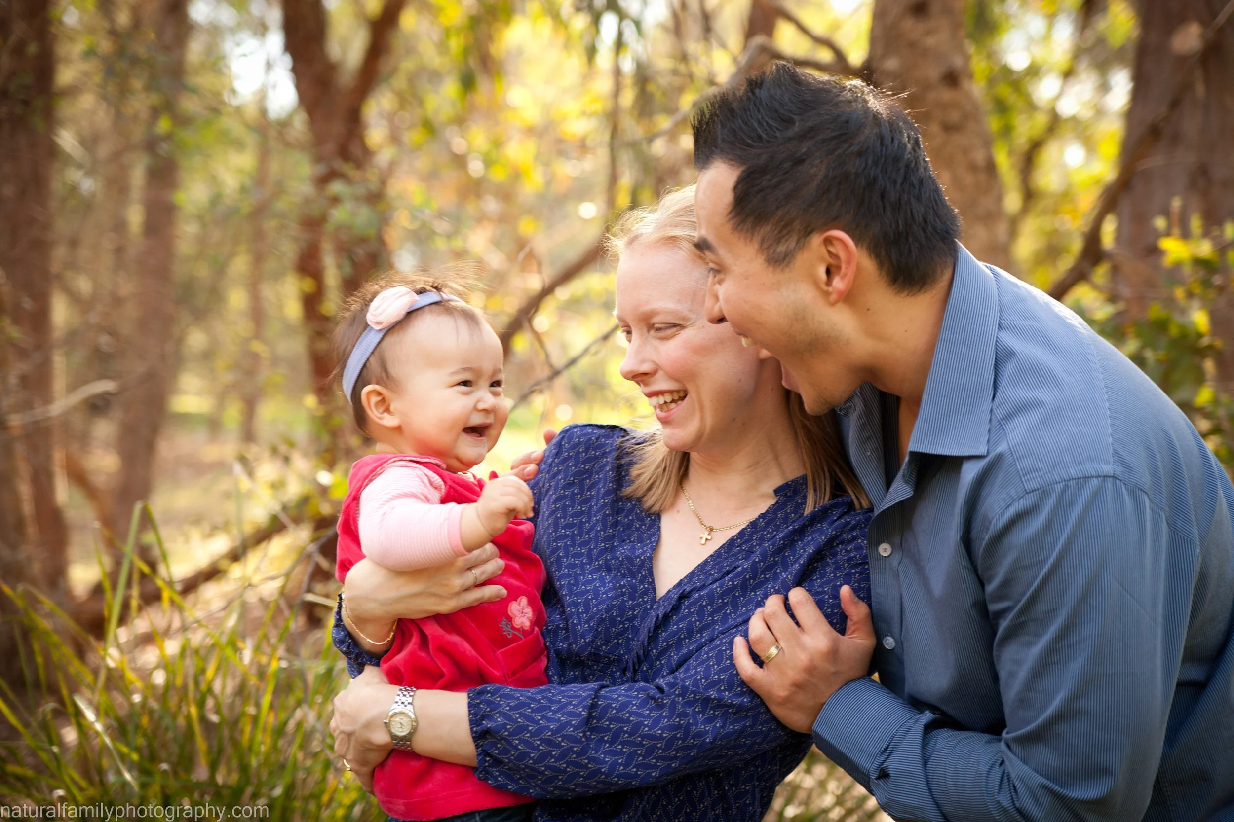 A family of three, consisting of a mother, father, and a baby girl, in a forest setting during autumn, smiling and playing with each other.
