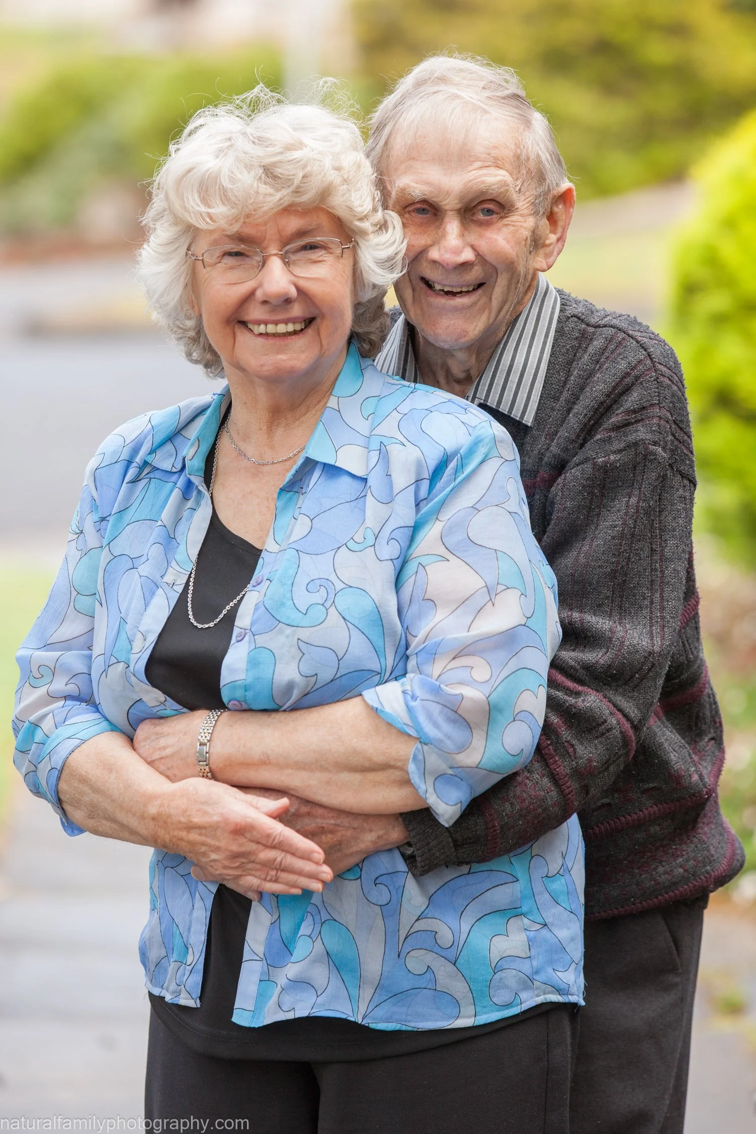 An elderly couple smiling outdoors with a blurred natural background, the woman wearing a blue patterned blouse and the man in a dark sweater.