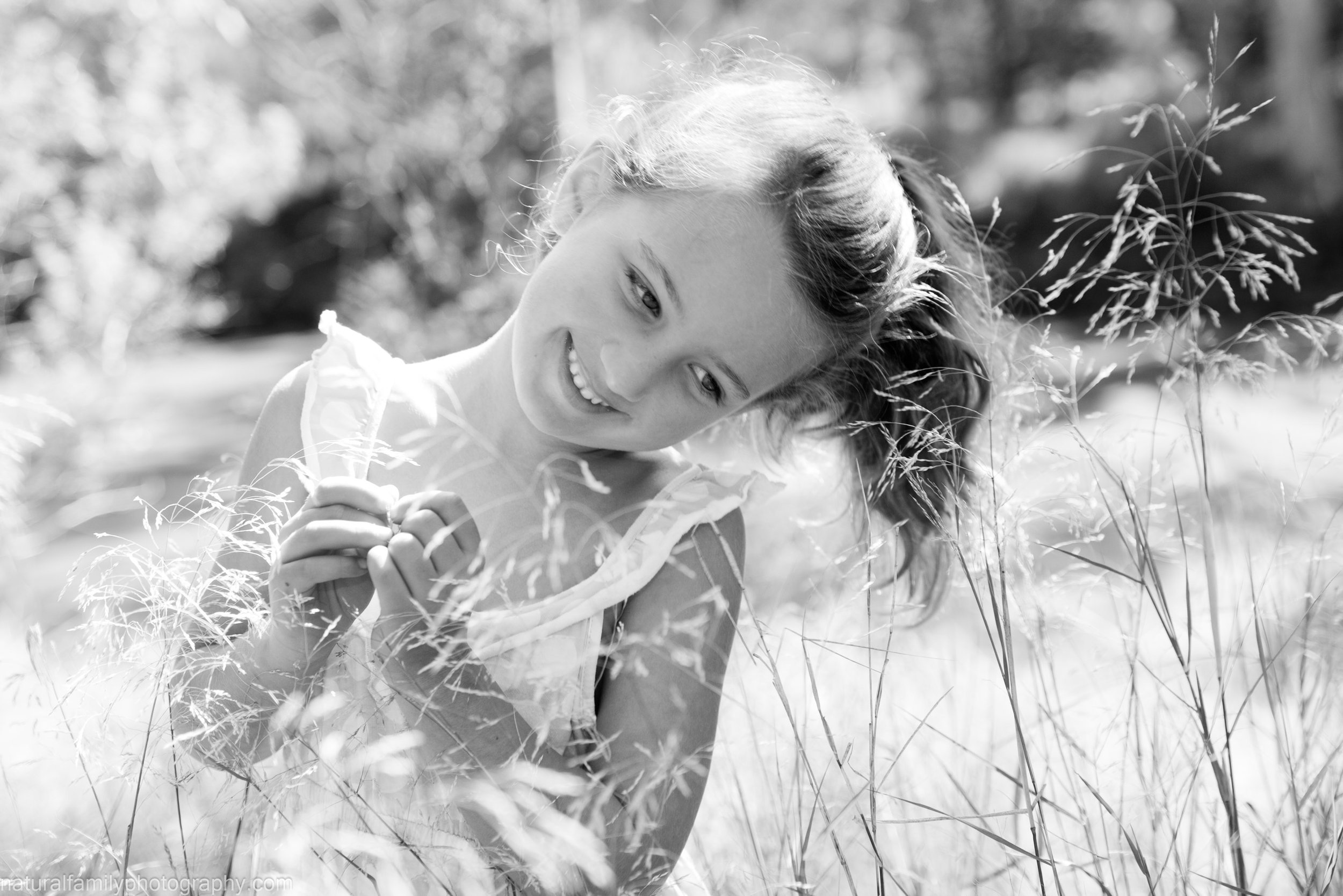 Smiling young woman outdoors in a field of tall grass, with sunlight and trees in the background, captured in black and white.