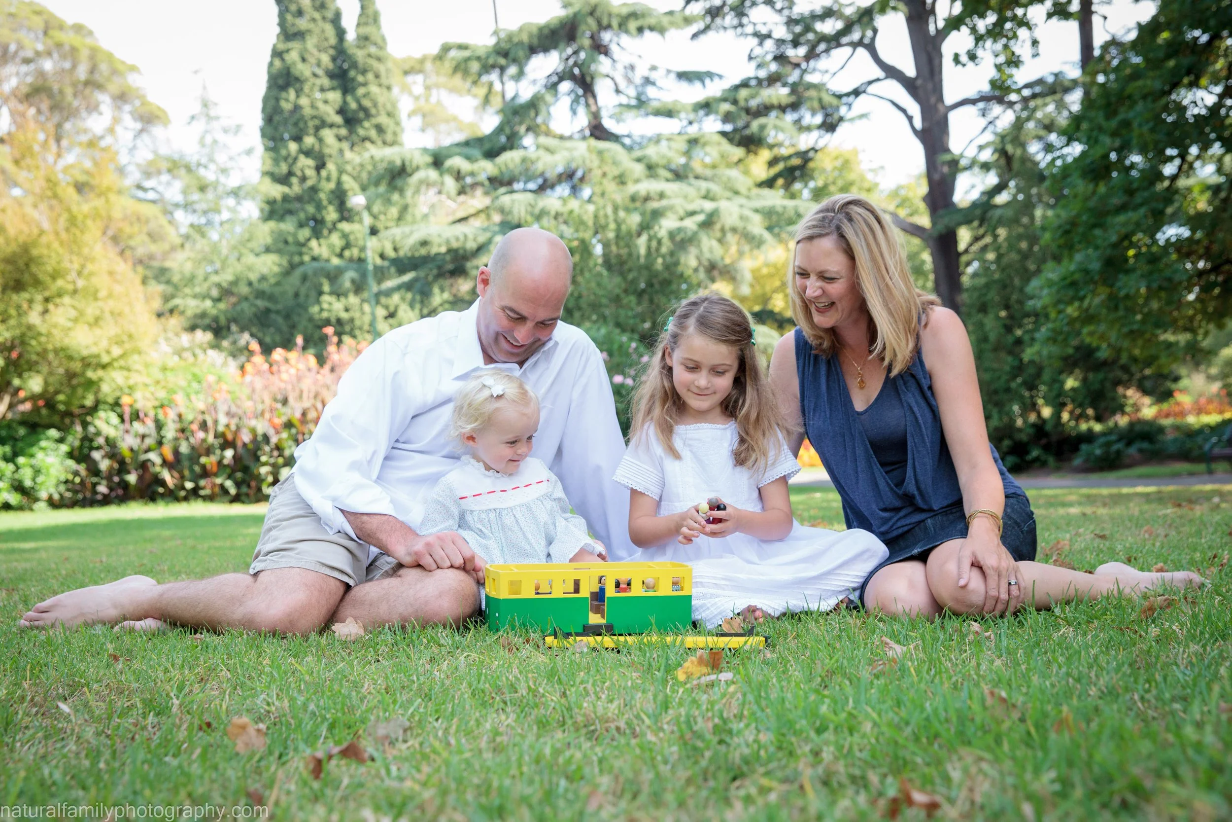 Family of four sitting on the grass in a park, playing with a yellow and green toy set, surrounded by trees and colorful plants on a sunny day.