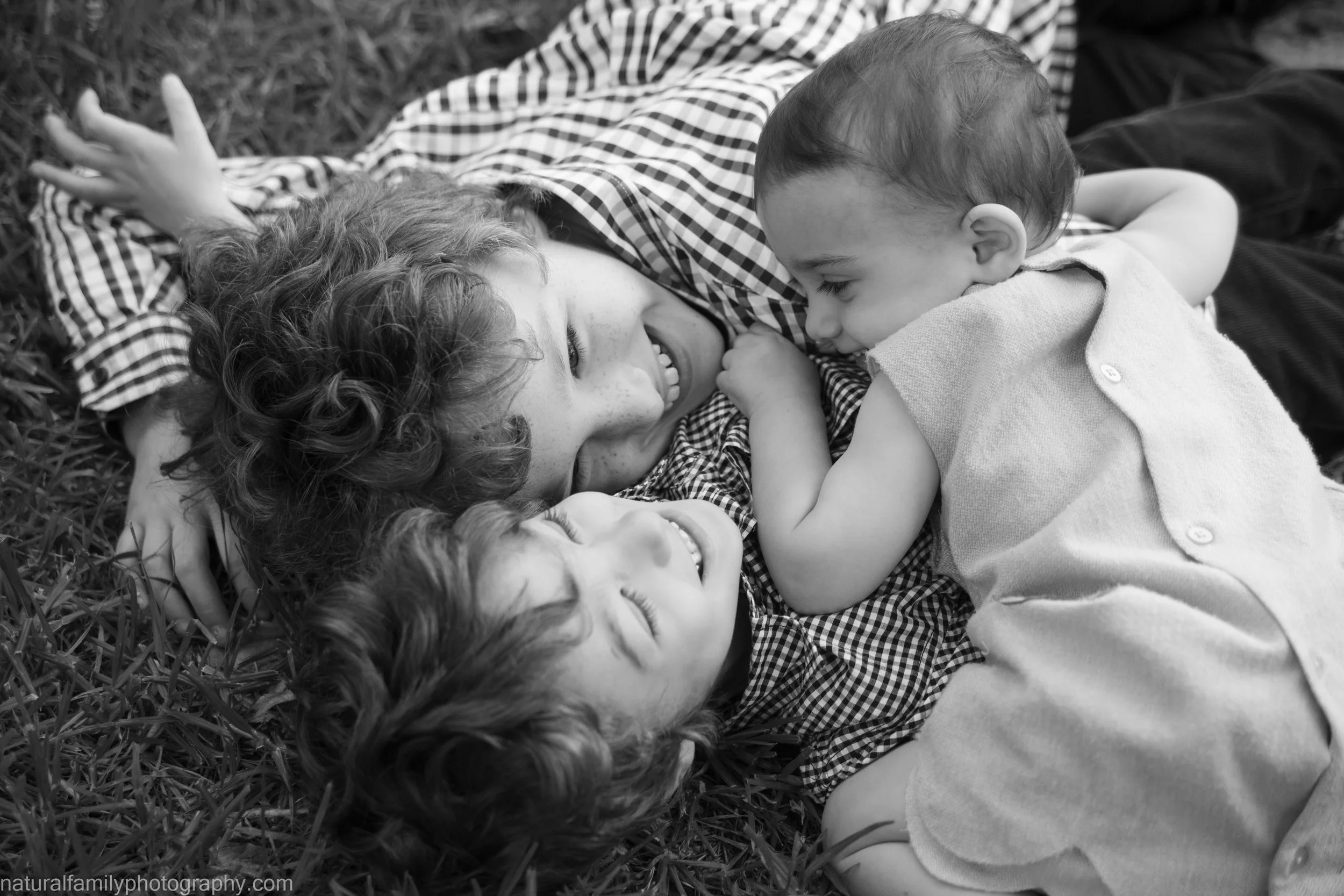 Three children lying on grass, smiling and playing together.