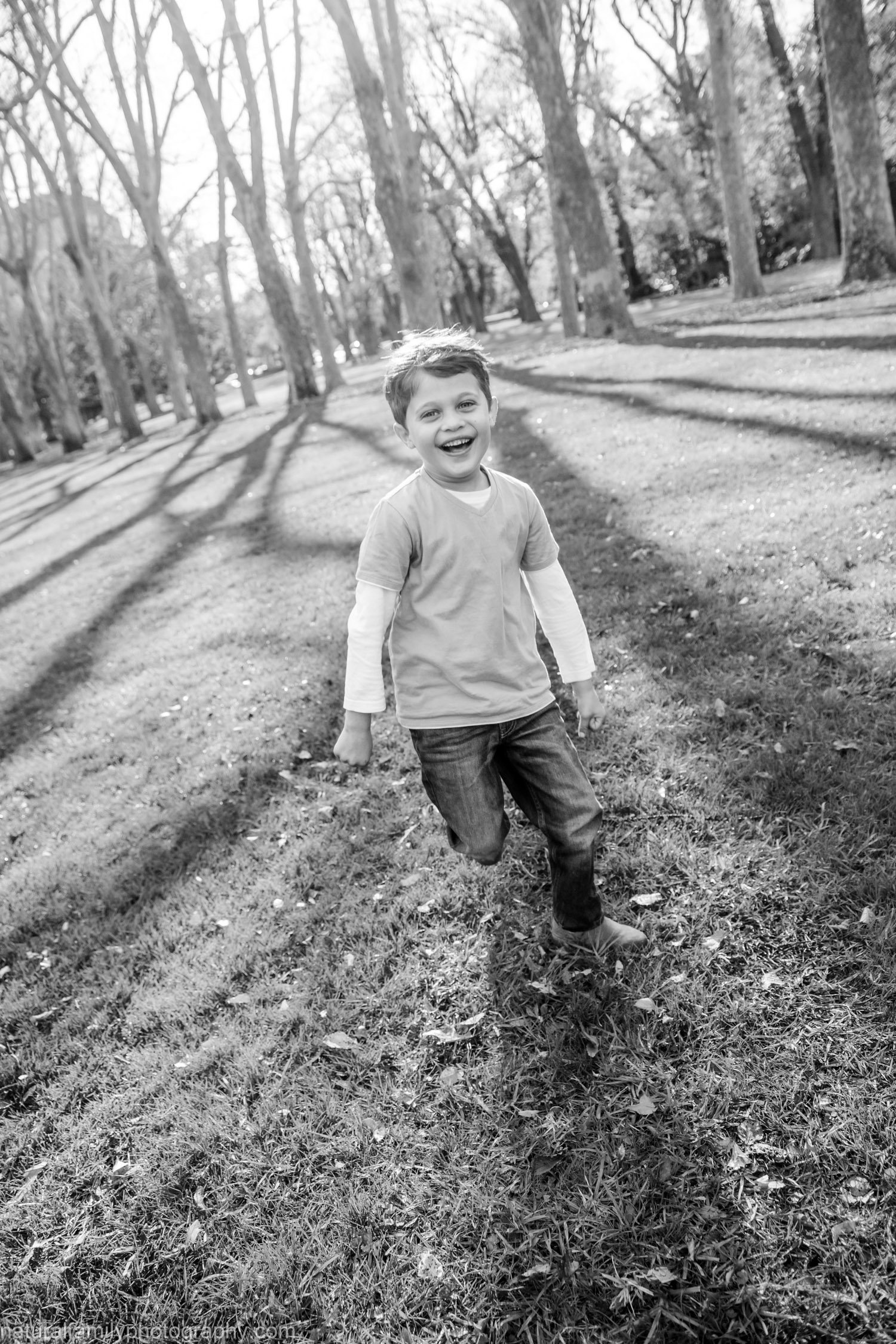 A young boy running on grass in a park with trees, smiling and enjoying himself.
