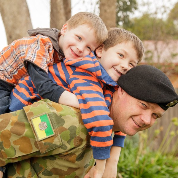 A soldier in camouflage uniform giving his two young sons a piggyback ride outdoors during daytime. Heartfelt portraiture by Natural Family Photography, Melbourne.