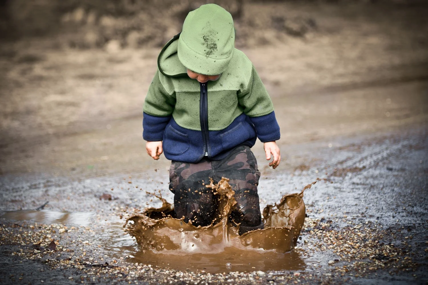 Young child wearing green and blue jacket and camouflage pants playing in muddy puddle outdoors.