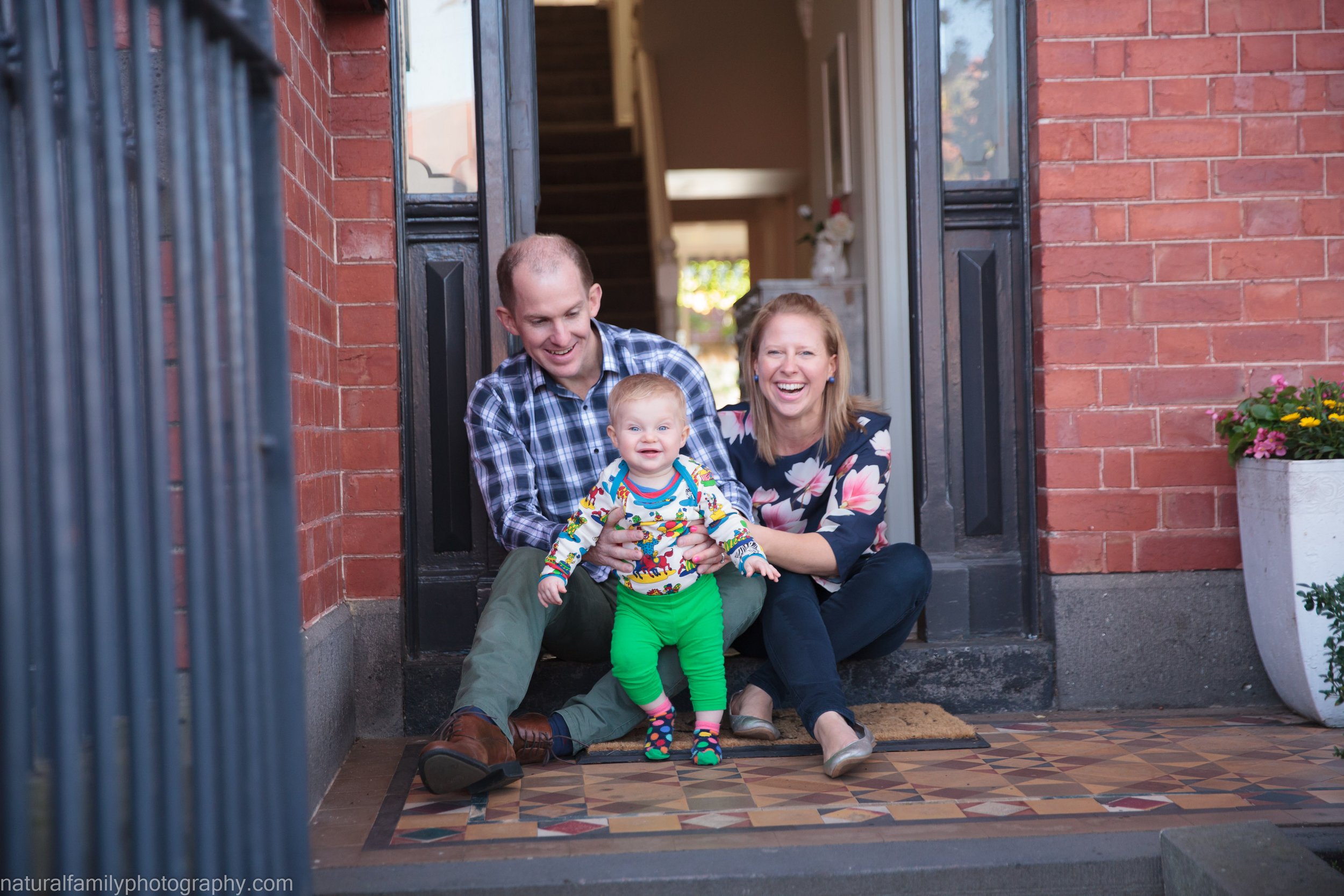 A family sitting on the front porch of their home, smiling and holding a young child, with a brick house and potted flowers visible.