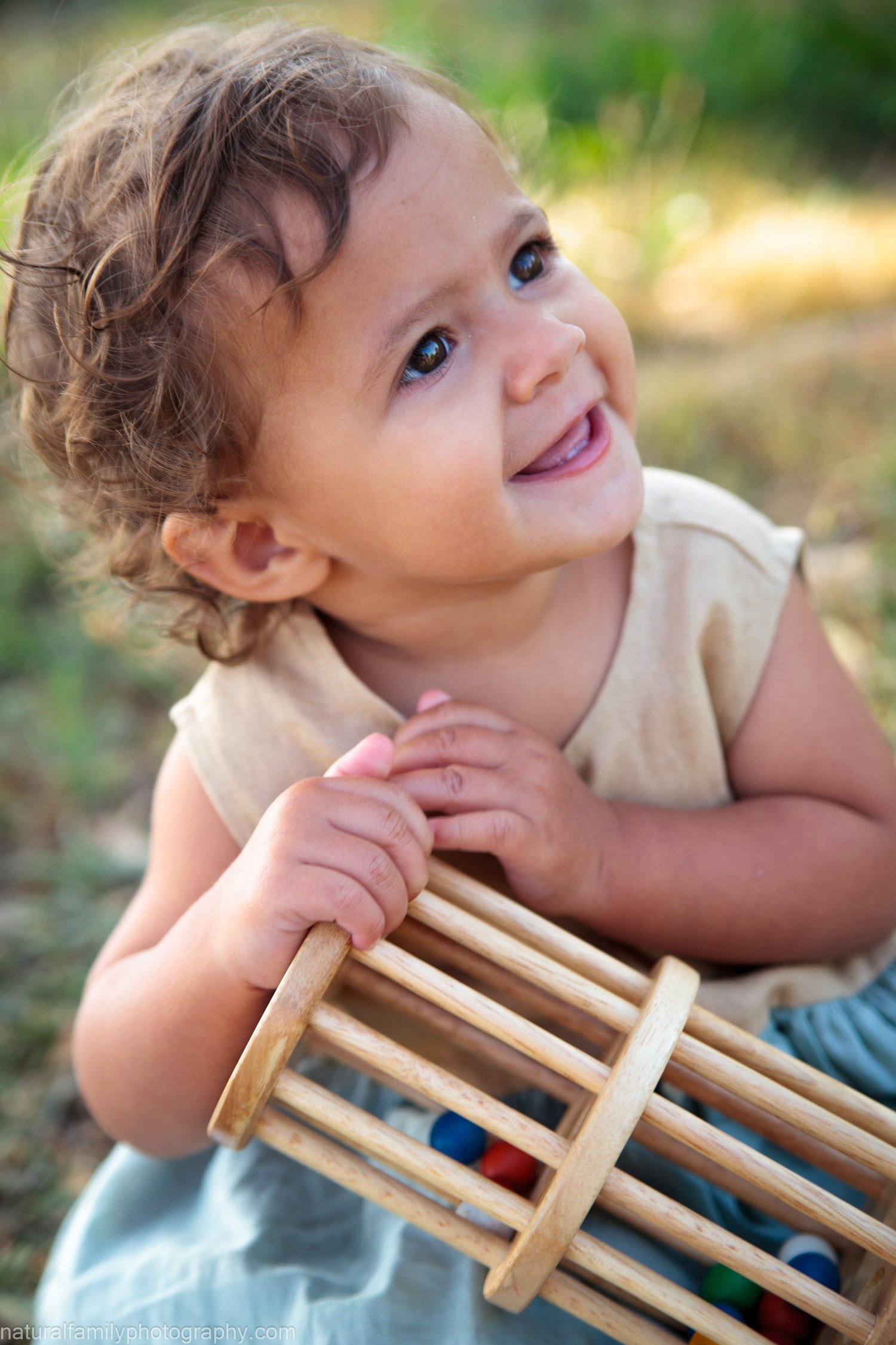 A young child with curly hair and bright eyes holding a small wooden toy with colorful balls outdoors, smiling and looking upward. Playful, joyful portrait by Natural Family Photography, Melbourne.