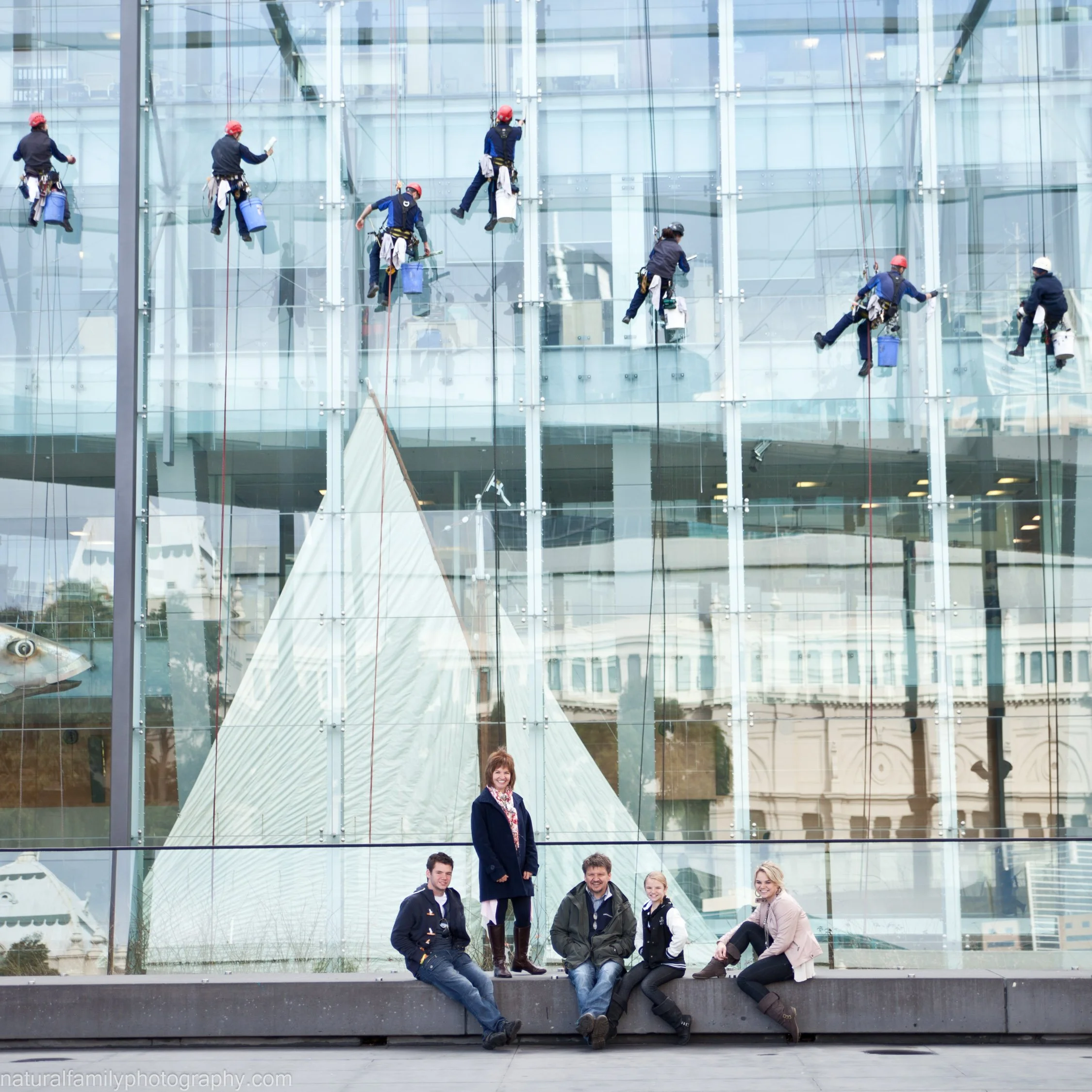 A glass building with cleaning workers wearing red and white helmets cleaning the windows while suspended by ropes. In front, four people are sitting on a ledge, and one woman is standing behind them, smiling.
