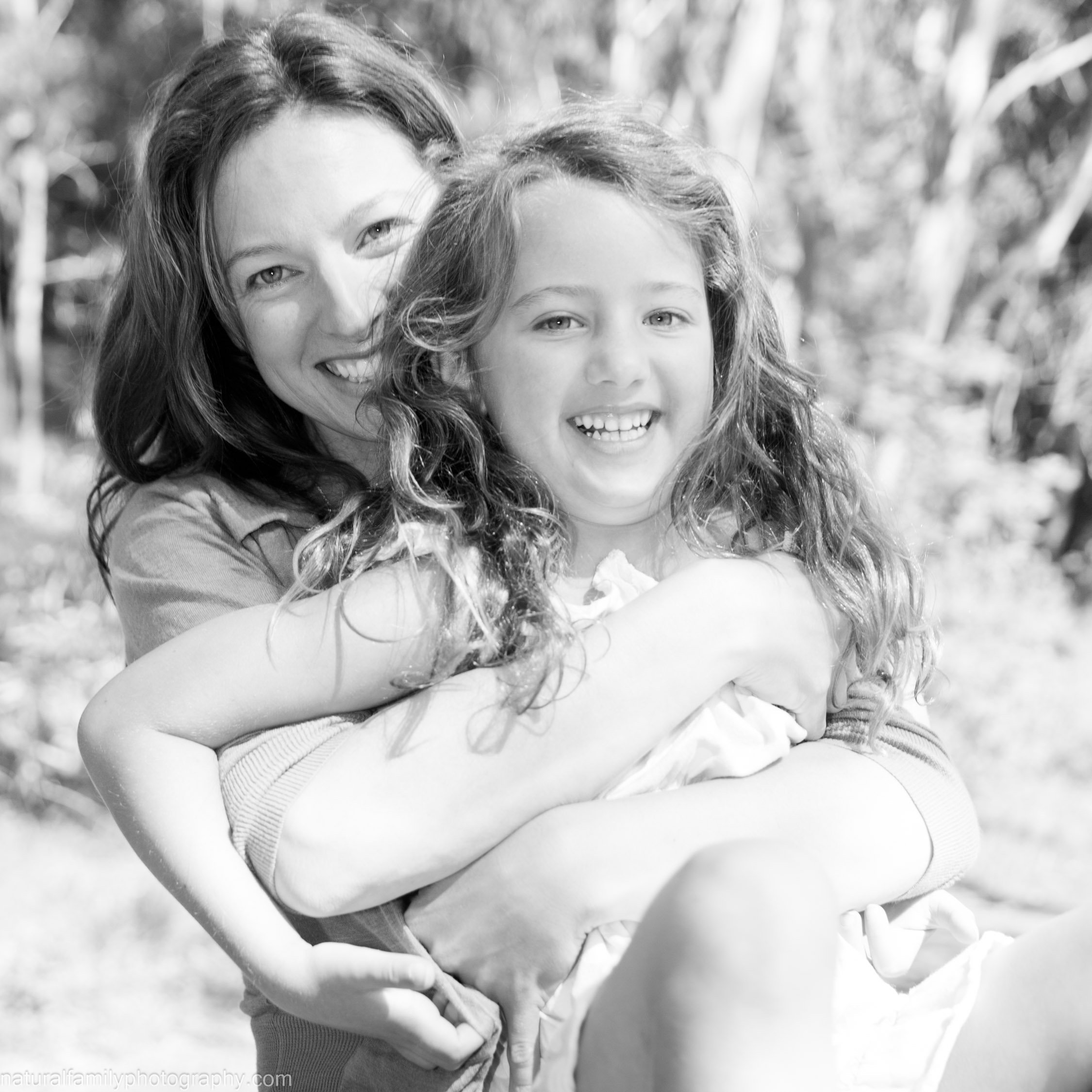 A woman and a young girl smiling and hugging outdoors in a natural setting. Classic black and white portrait by Natural Family Photography, Melbourne.