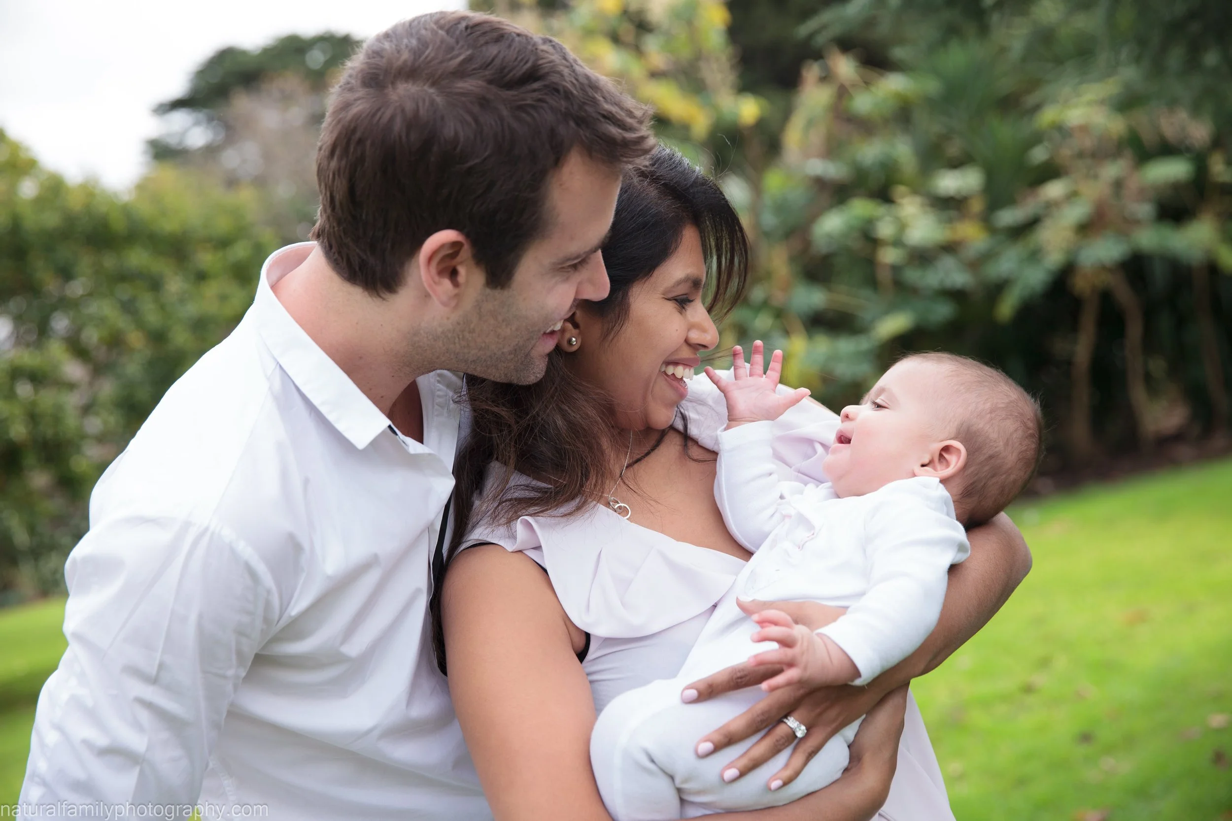 A happy family of three outdoors, with a woman holding a baby and a man leaning in, all smiling and looking at each other.
