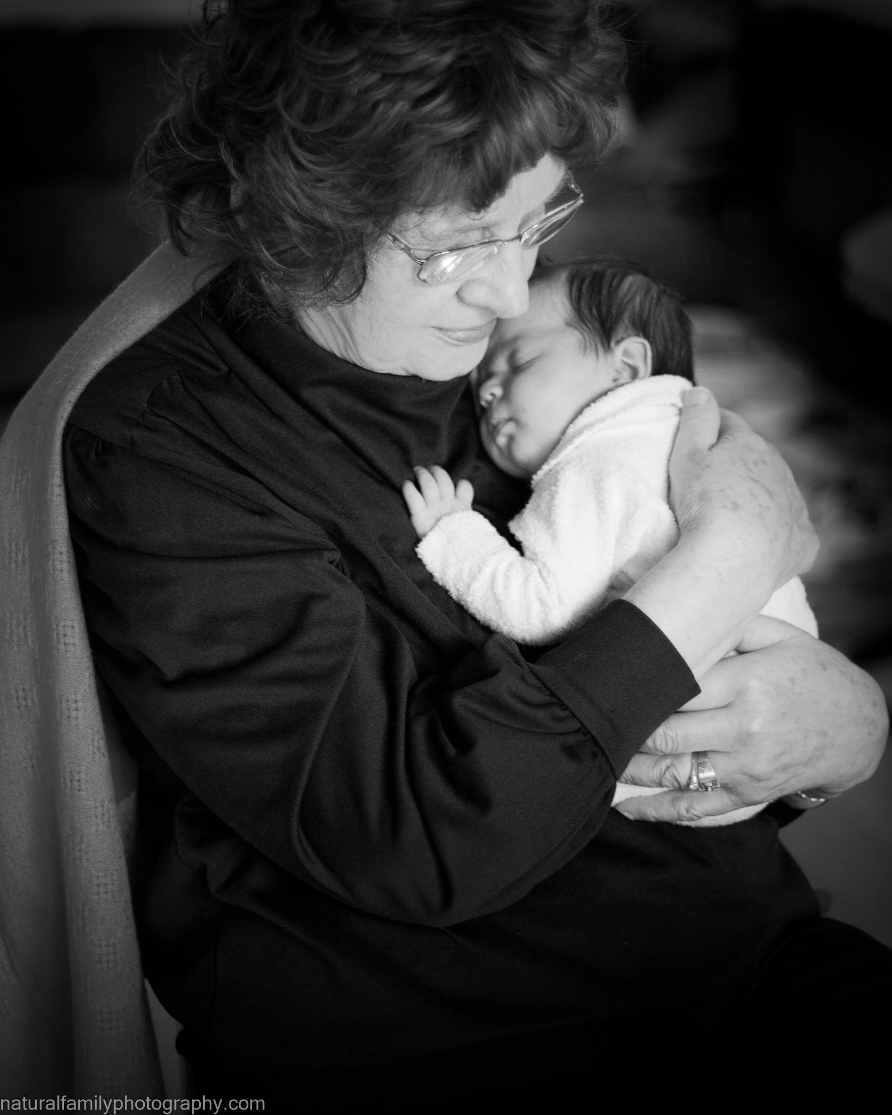 A woman holding a sleeping baby close to her chest, smiling gently in a black-and-white photo. Generational portraiture by Natural Family Photography, Melbourne.