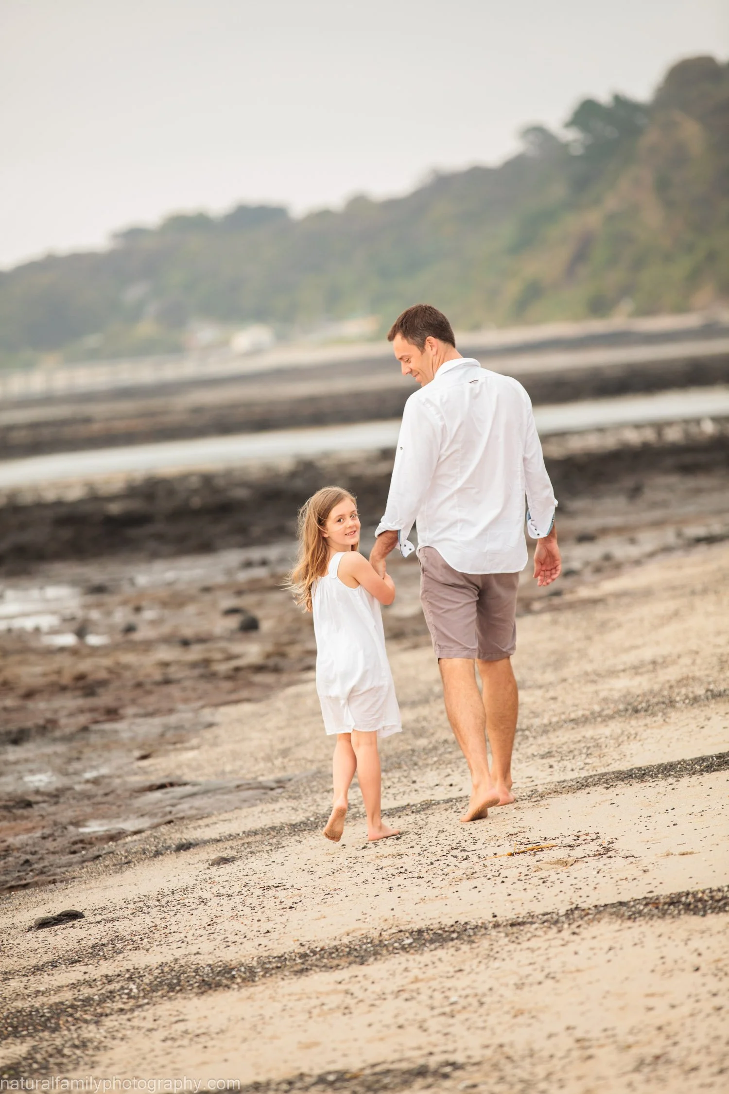 A man and girl holding hands walking on a sandy beach, with the girl looking back and smiling at the camera, and they are dressed in white summer clothing.