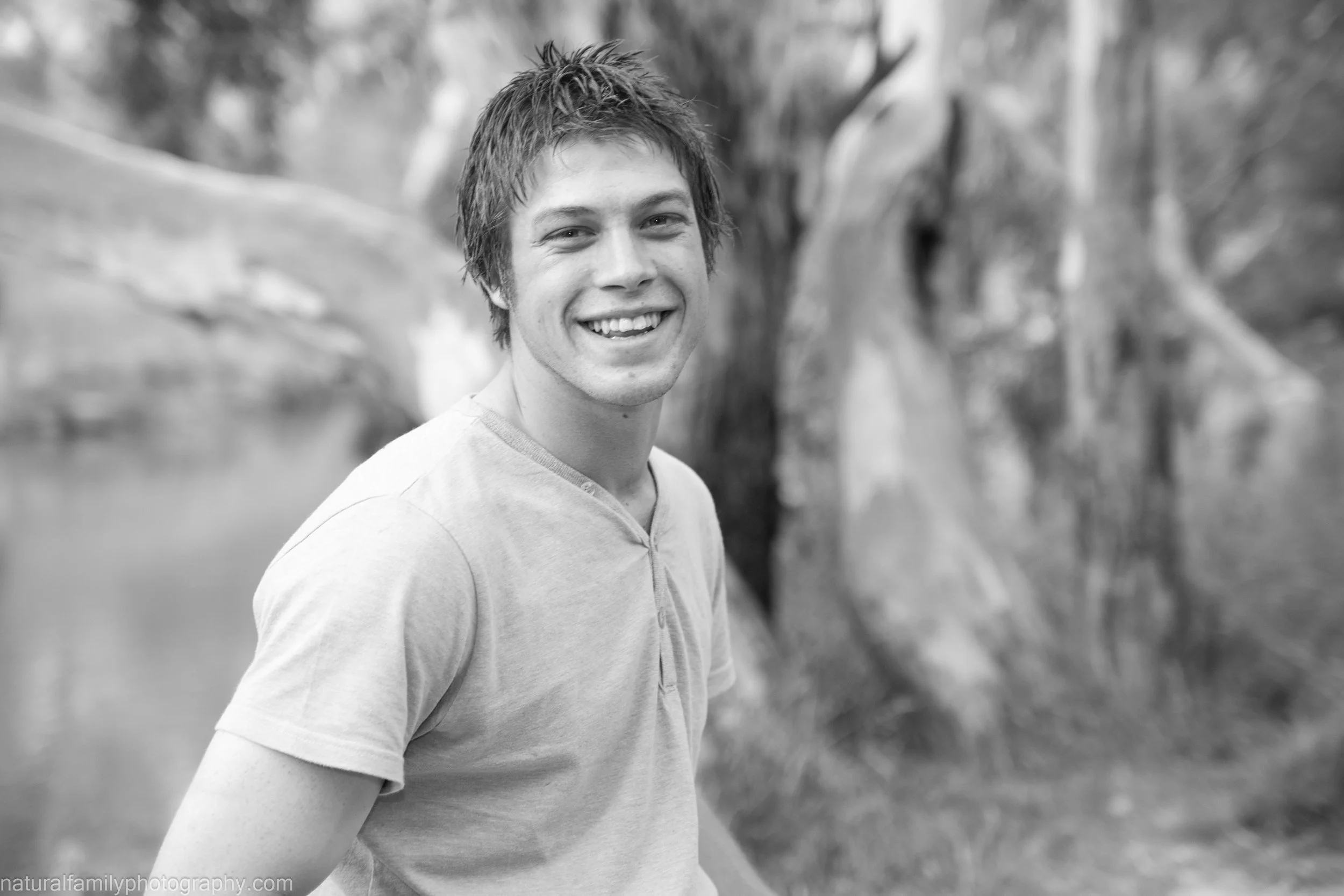 Black and white photo of a smiling young man standing outdoors in front of trees.