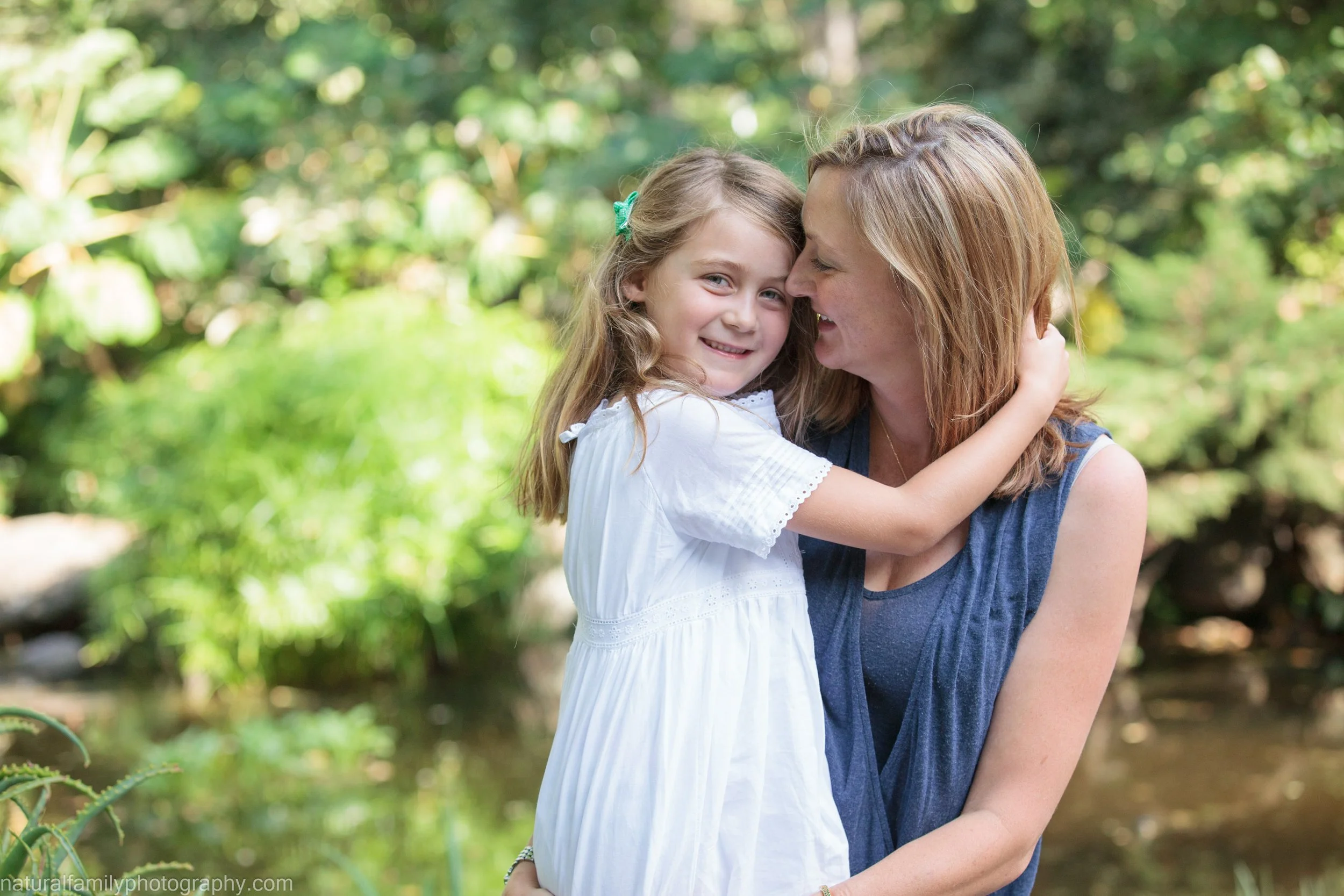 A woman and a young girl embracing outdoors near a stream, smiling happily at each other.