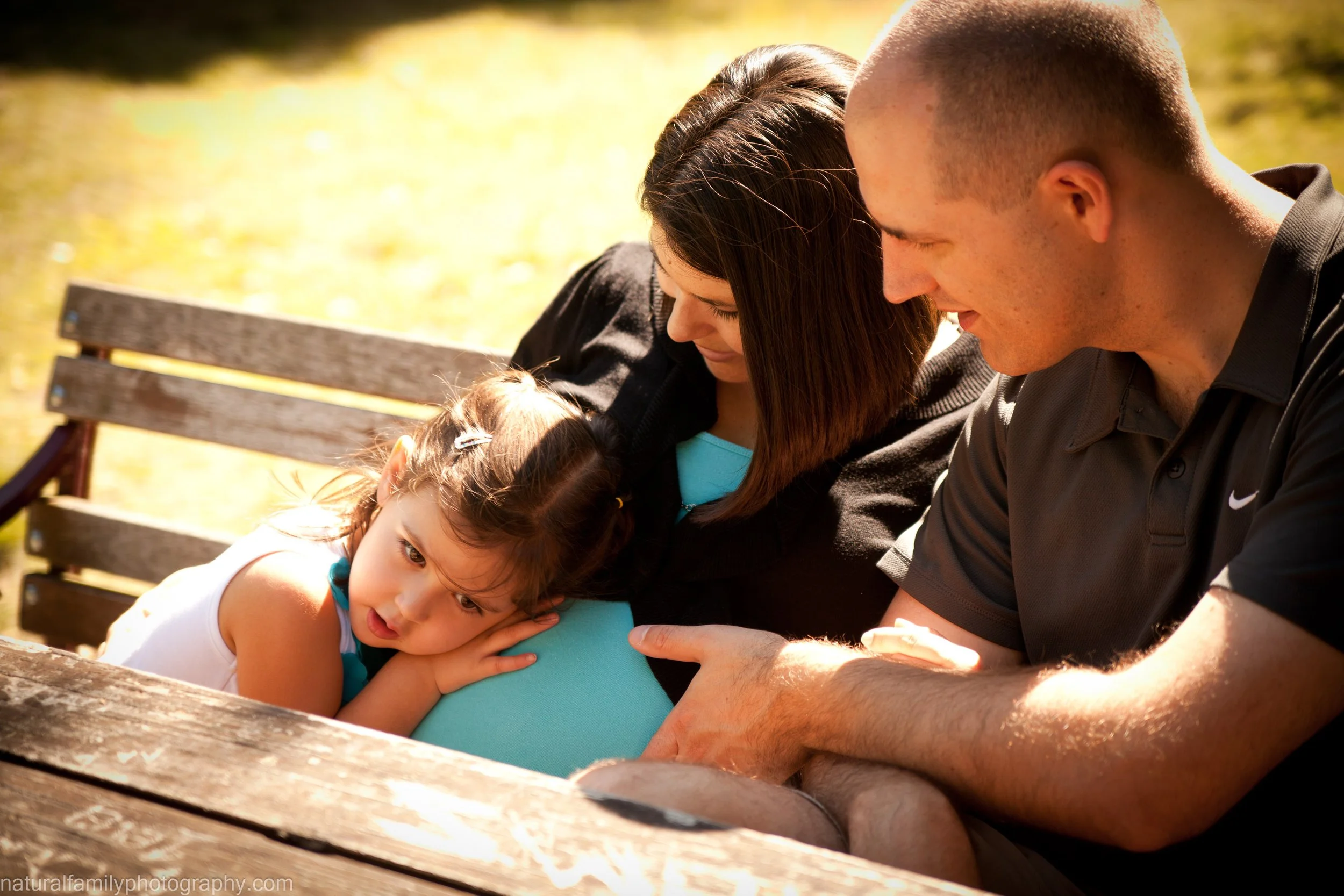 A family of three sitting outdoors on a wooden bench, with the young girl resting her head on her mother's lap and the father smiling as he touches her belly, enjoying a sunny day.