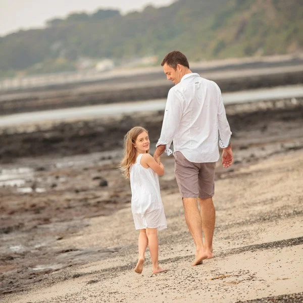 A man and young girl walking barefoot on a beach with wet sand and rocks, holding hands, with a hilly landscape in the background. Beach portraiture by Natural Family Photography, Melbourne.