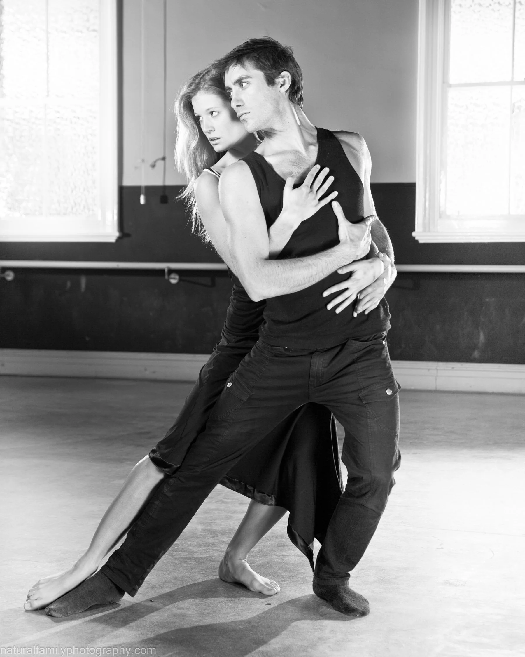 Two dancers practicing a slow dance or ballet in a studio, with light coming through the windows, black and white photo. Couples portrait by Natural Family Photography, Melbourne.