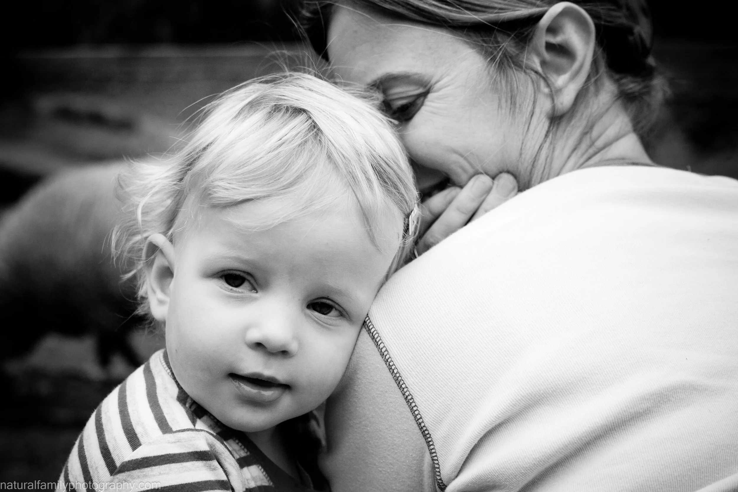 A young girl with light hair and a striped shirt is being hugged by her mother, who is smiling and has her eyes closed, with a blurry background. Classic black and white photography by Natural Family Photography, Melbourne.