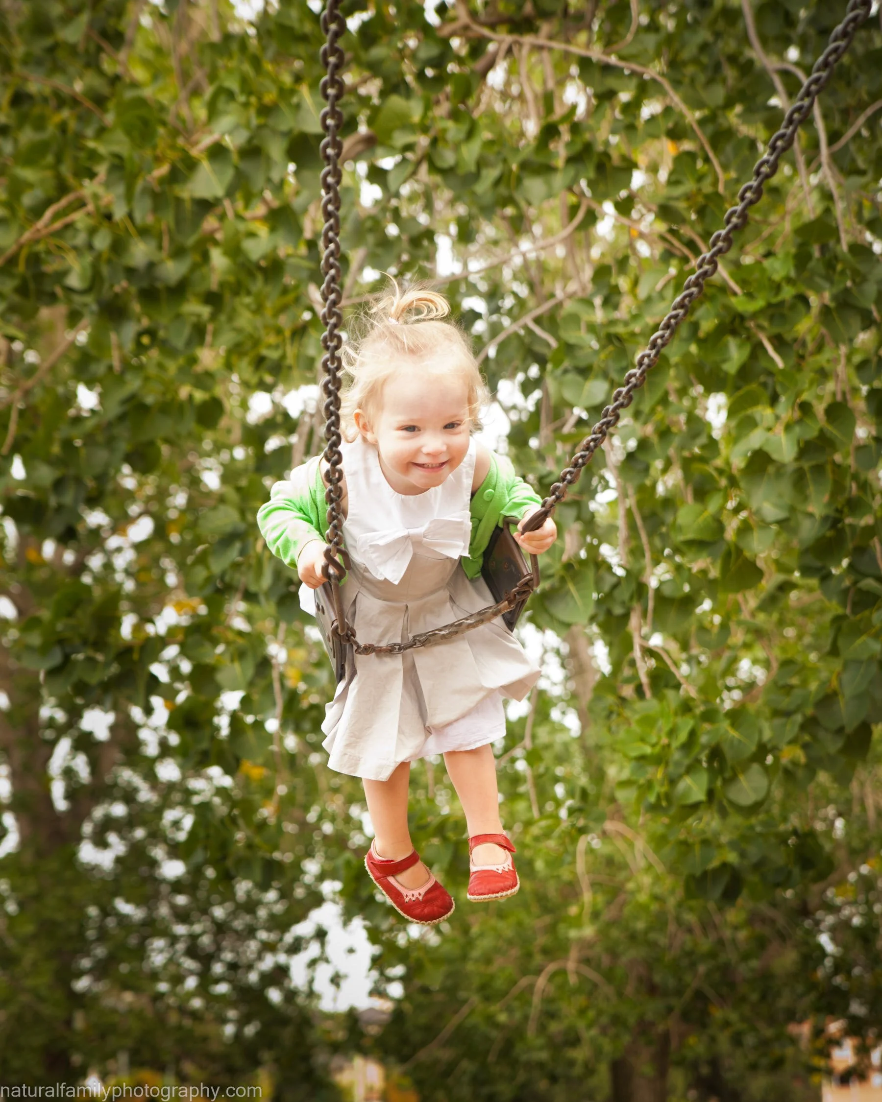A young girl with blonde hair in a ponytail smiling as she pushes herself on a swing set in a park, surrounded by green trees. Portrait by Natural Family Photography, Melbourne.