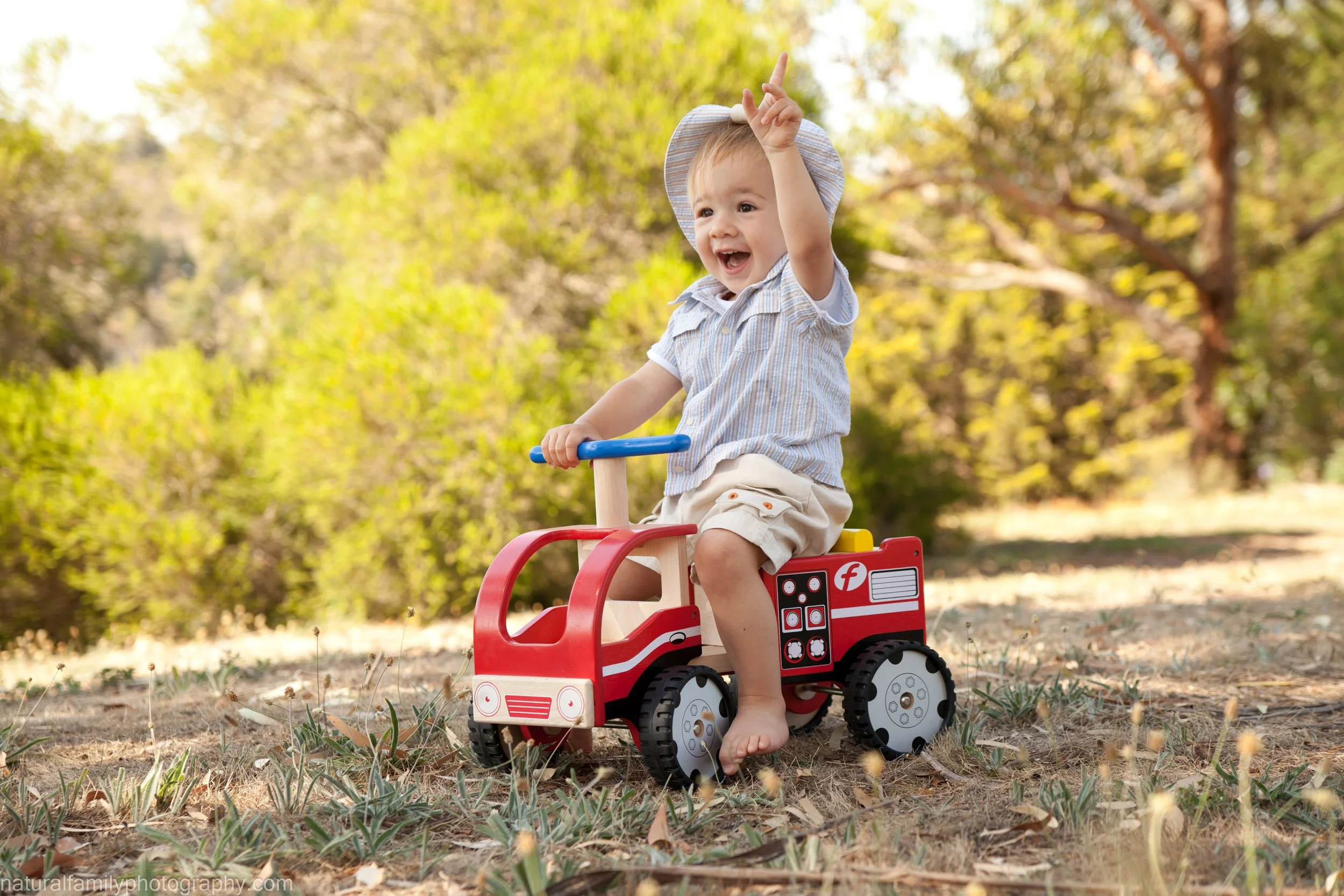 A young child riding a red toy fire truck outdoors in a park-like setting, smiling and raising one hand. Portrait by Natural Family Photography, Melbourne.