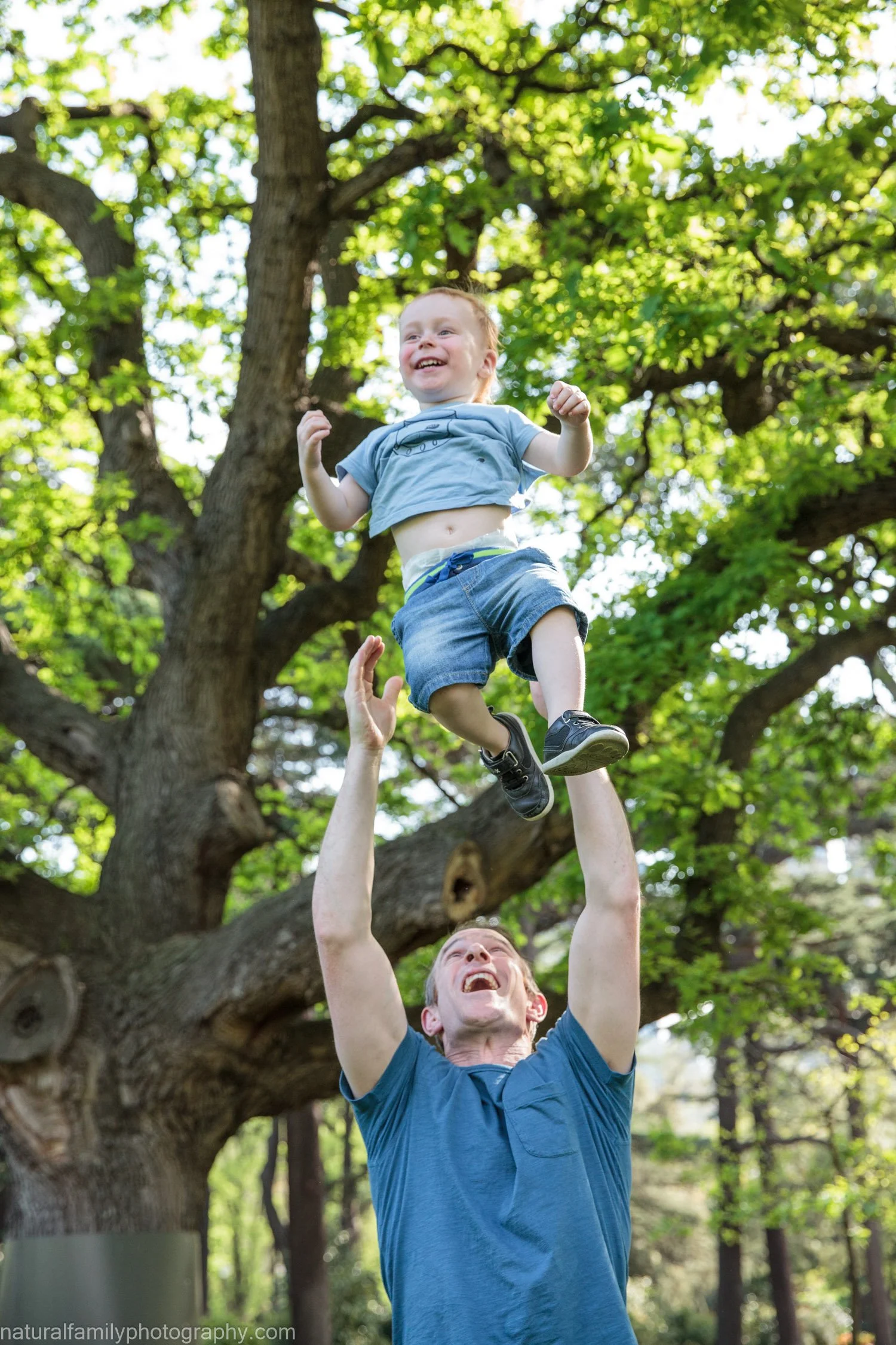 A man lifting a young boy in the air outdoors near a large tree with lush green leaves, smiling and enjoying a playful moment.