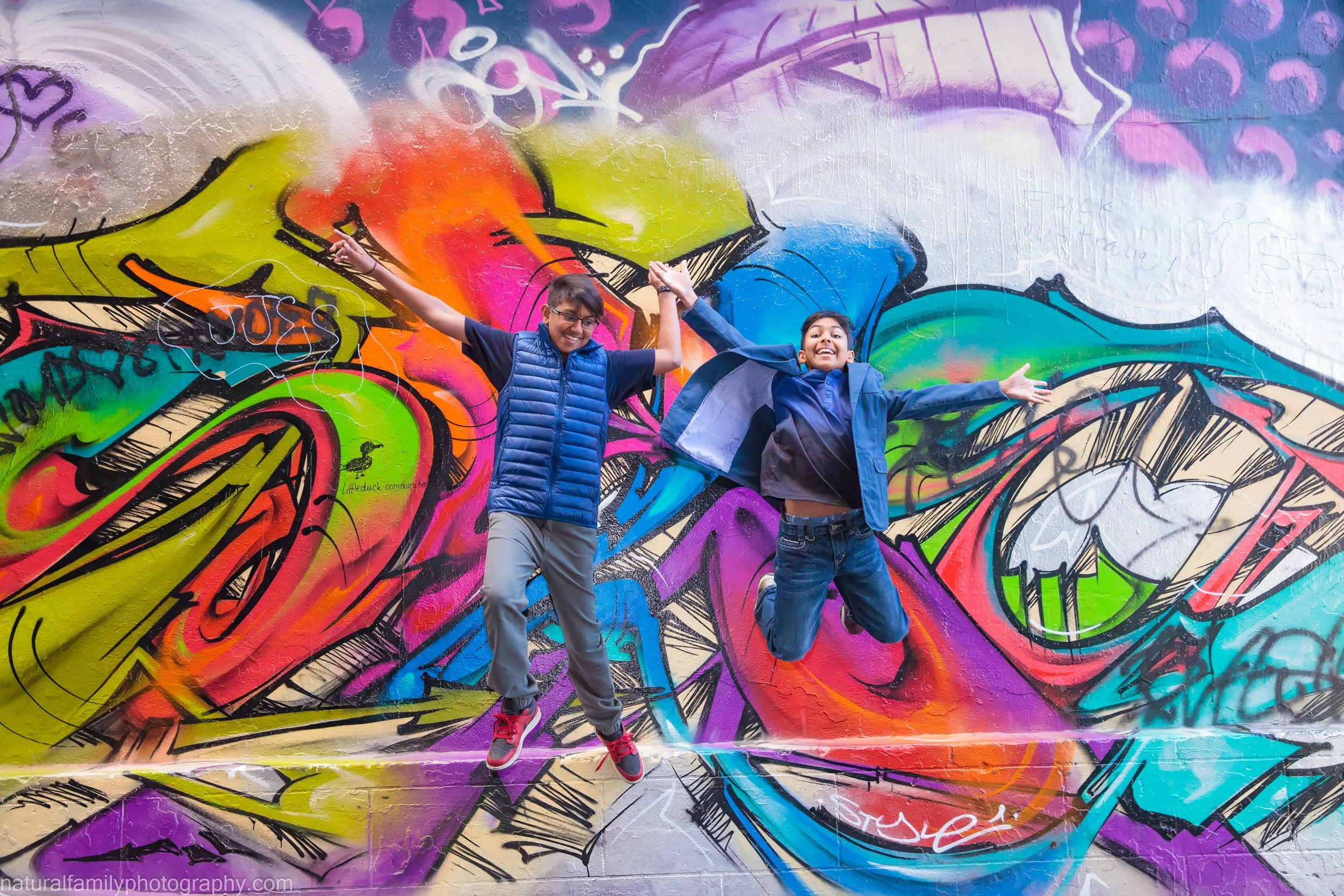 Two young girls jumping in front of a colorful graffiti mural on a wall, with arms raised and smiling.