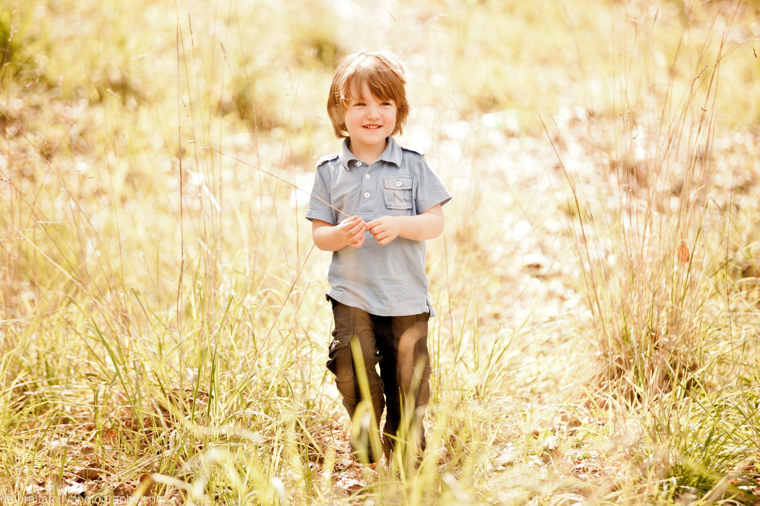 A young boy with brown hair and a wide smile walking through tall, golden grass outdoors on a sunny day.