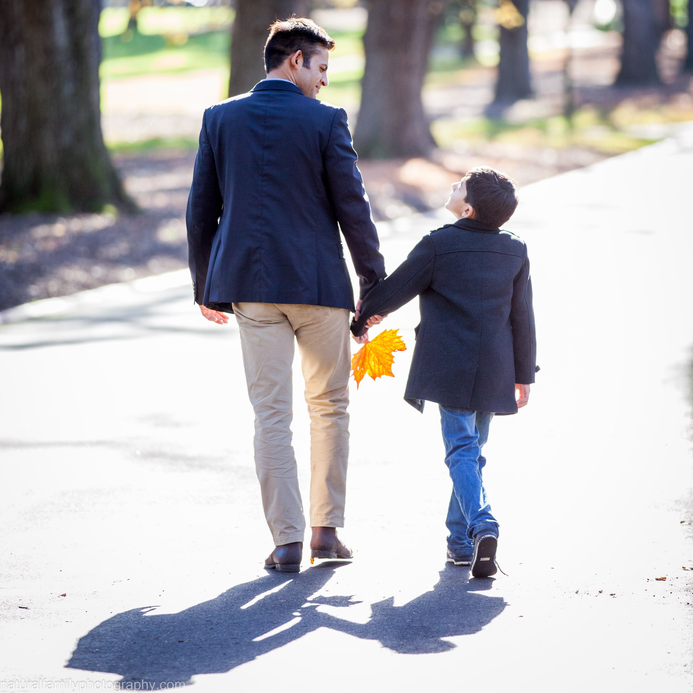 A man and a boy walking hand in hand on a tree-lined path, with the man holding some fall leaves.