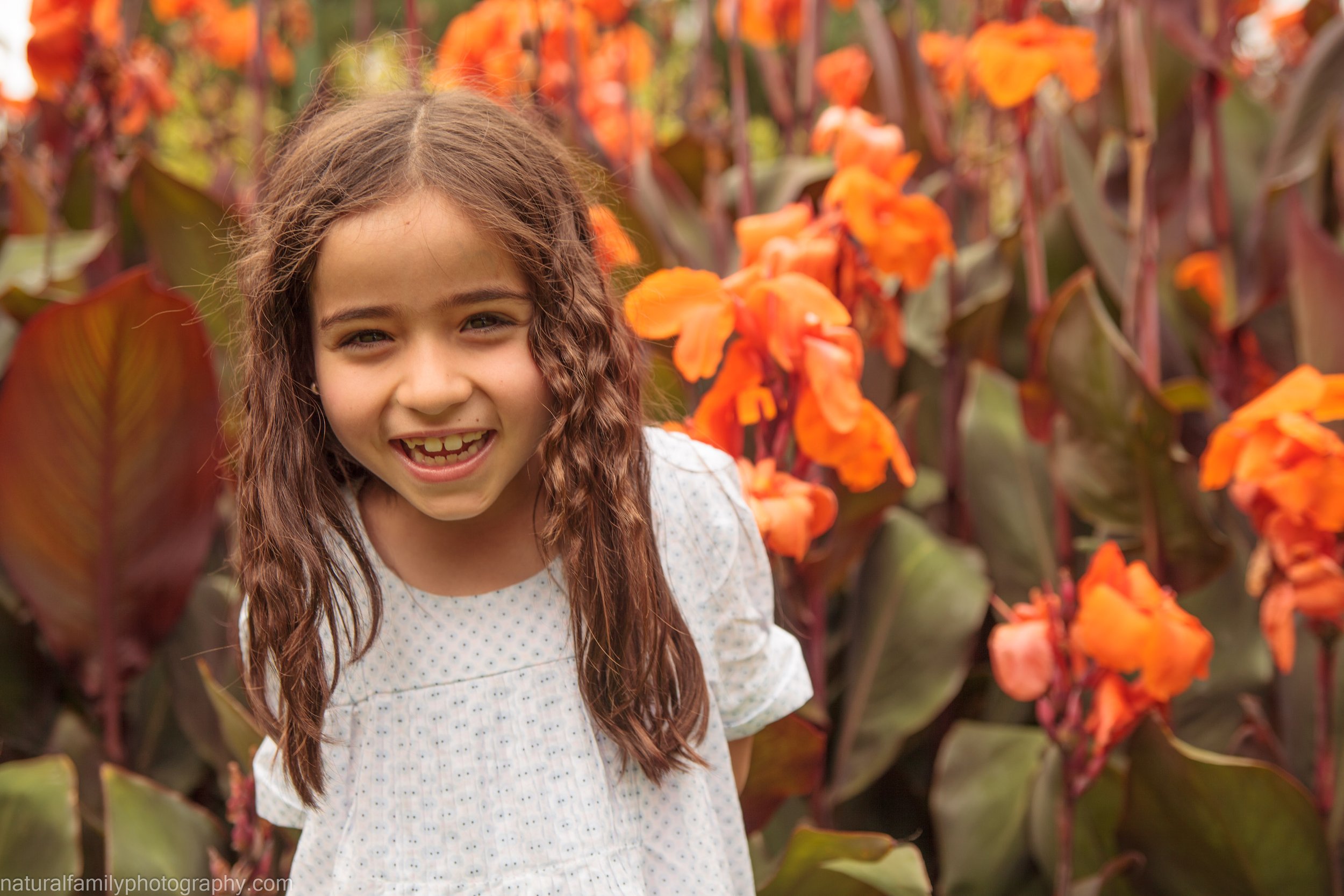 A young girl with long brown hair smiling in front of orange flowers in a garden.