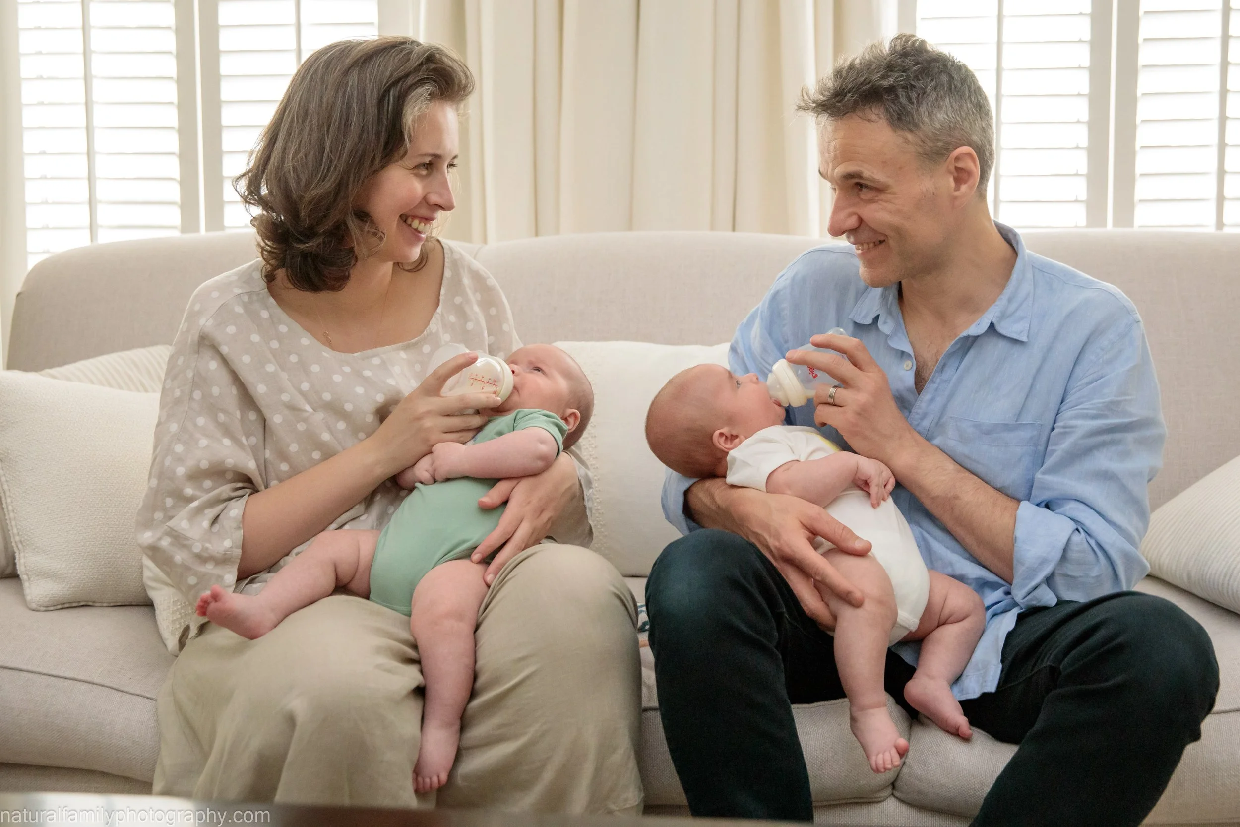 A couple of parents feeding their twin babies with bottles while sitting on a couch in a bright room.