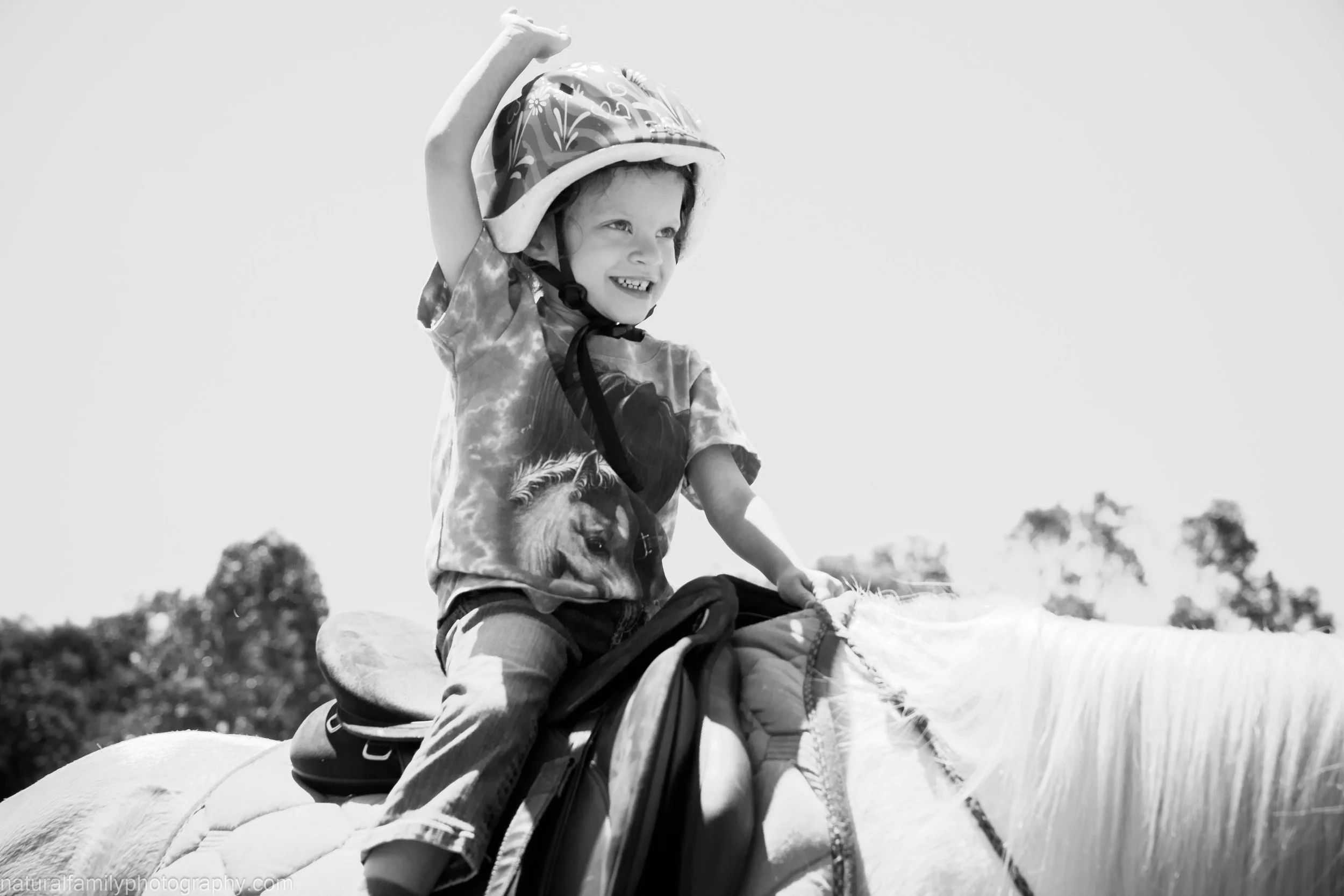 A young girl with a helmet riding a horse outdoors on a sunny day, smiling. Family and animal portraits by Natural Family Photography, Melbourne.