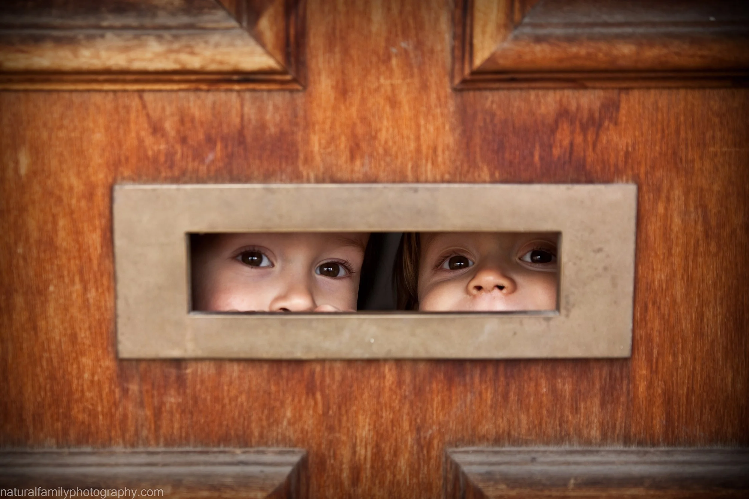 Two children peeking through a letter slot in a wooden door, with only their eyes and parts of their faces visible. Emotive, artistic portraiture by Natural Family Photography, Melbourne.