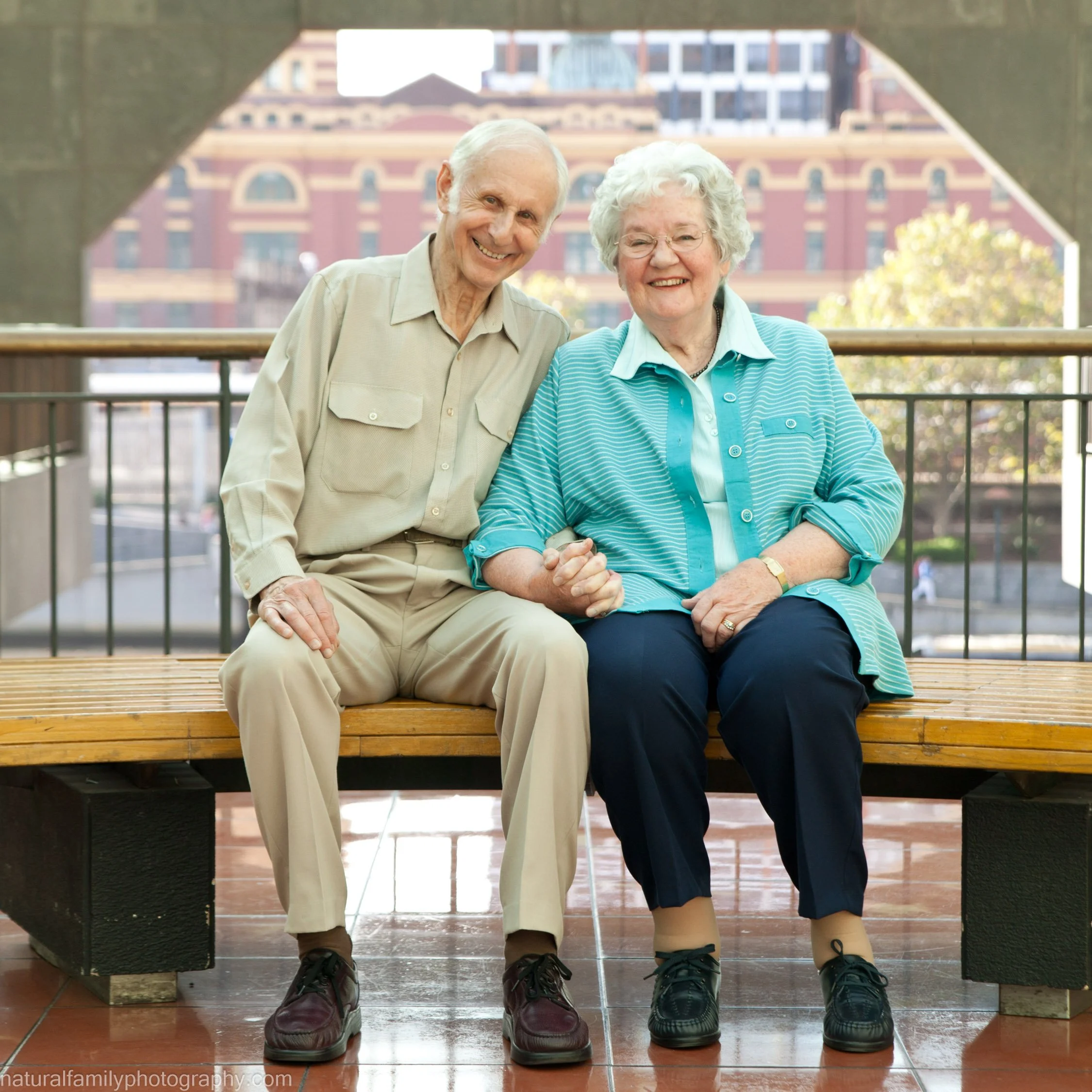 An elderly couple sitting on a wooden bench holding hands and smiling, with a cityscape in the background.