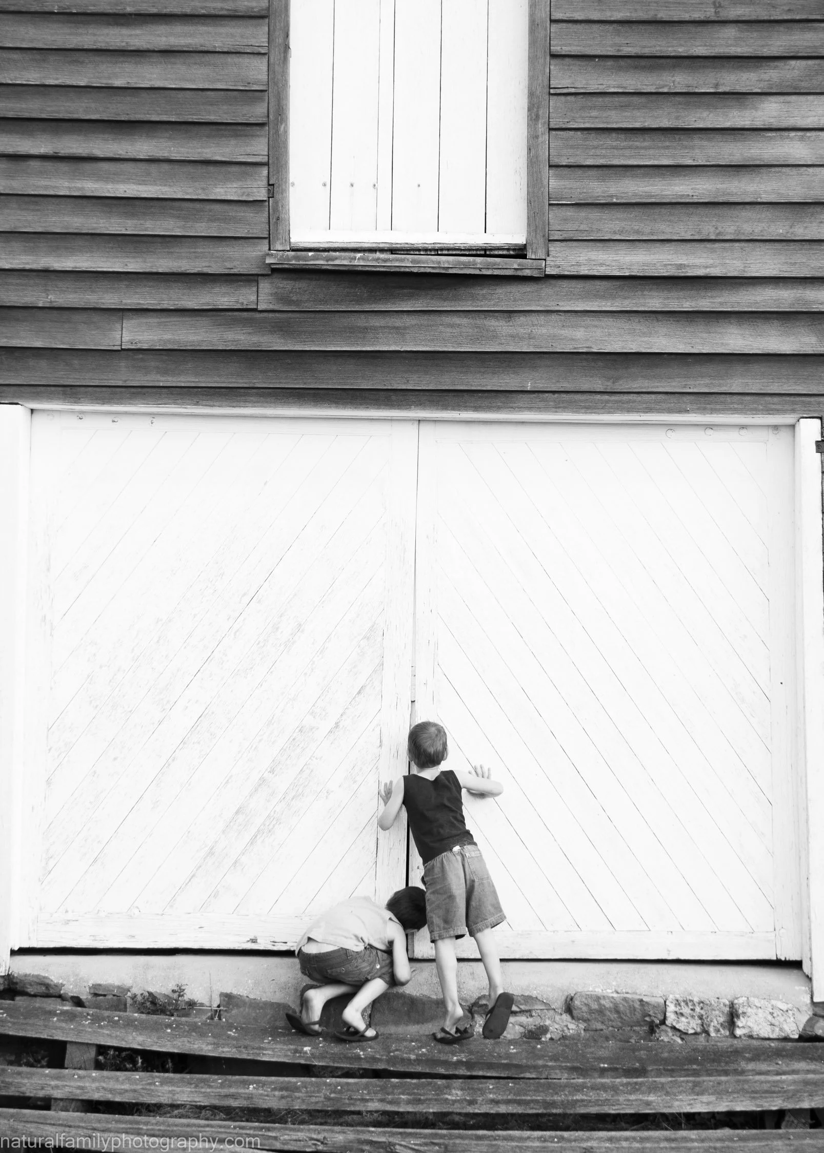 Two children playing in front of a large white barn door, with one child crouching and the other standing and peering inside.
