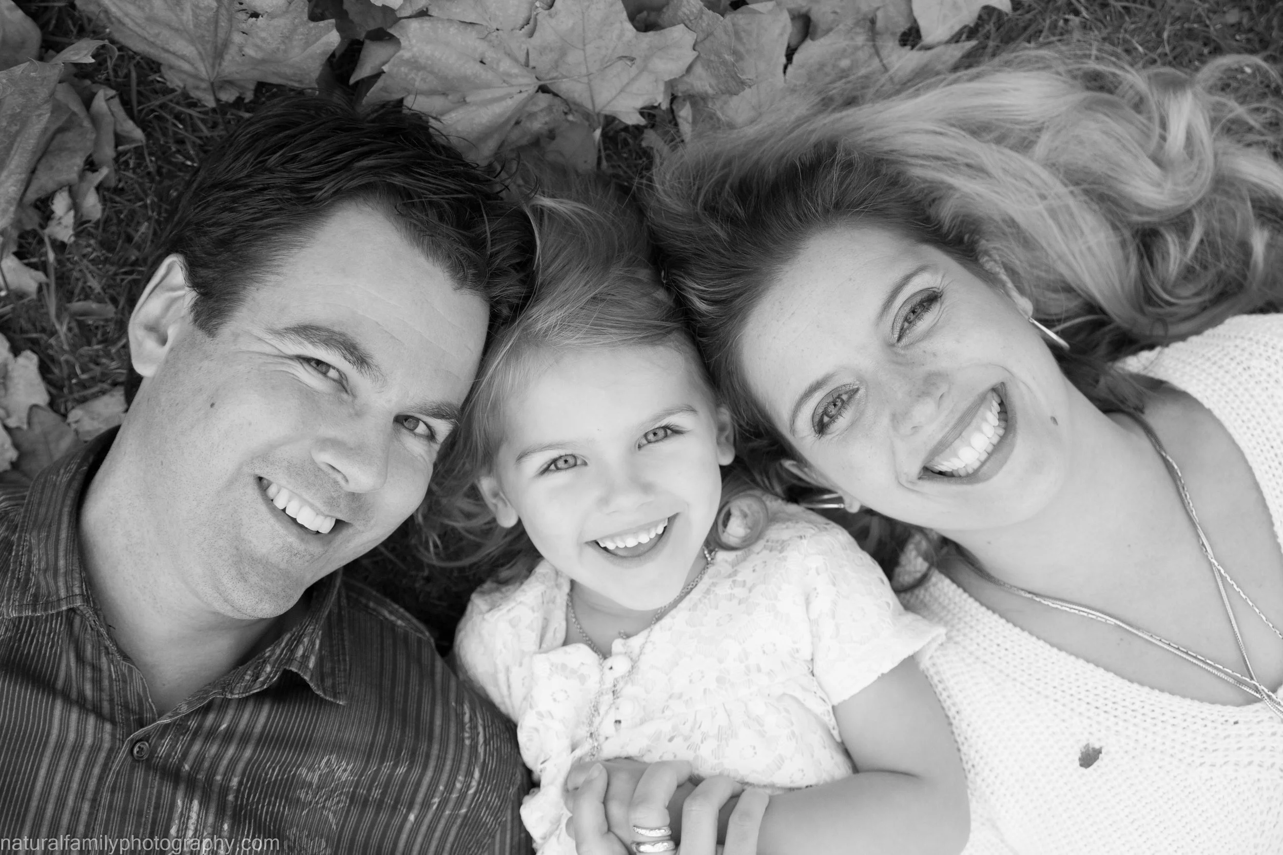 A happy family of three lying on the ground among fallen leaves, smiling at the camera.