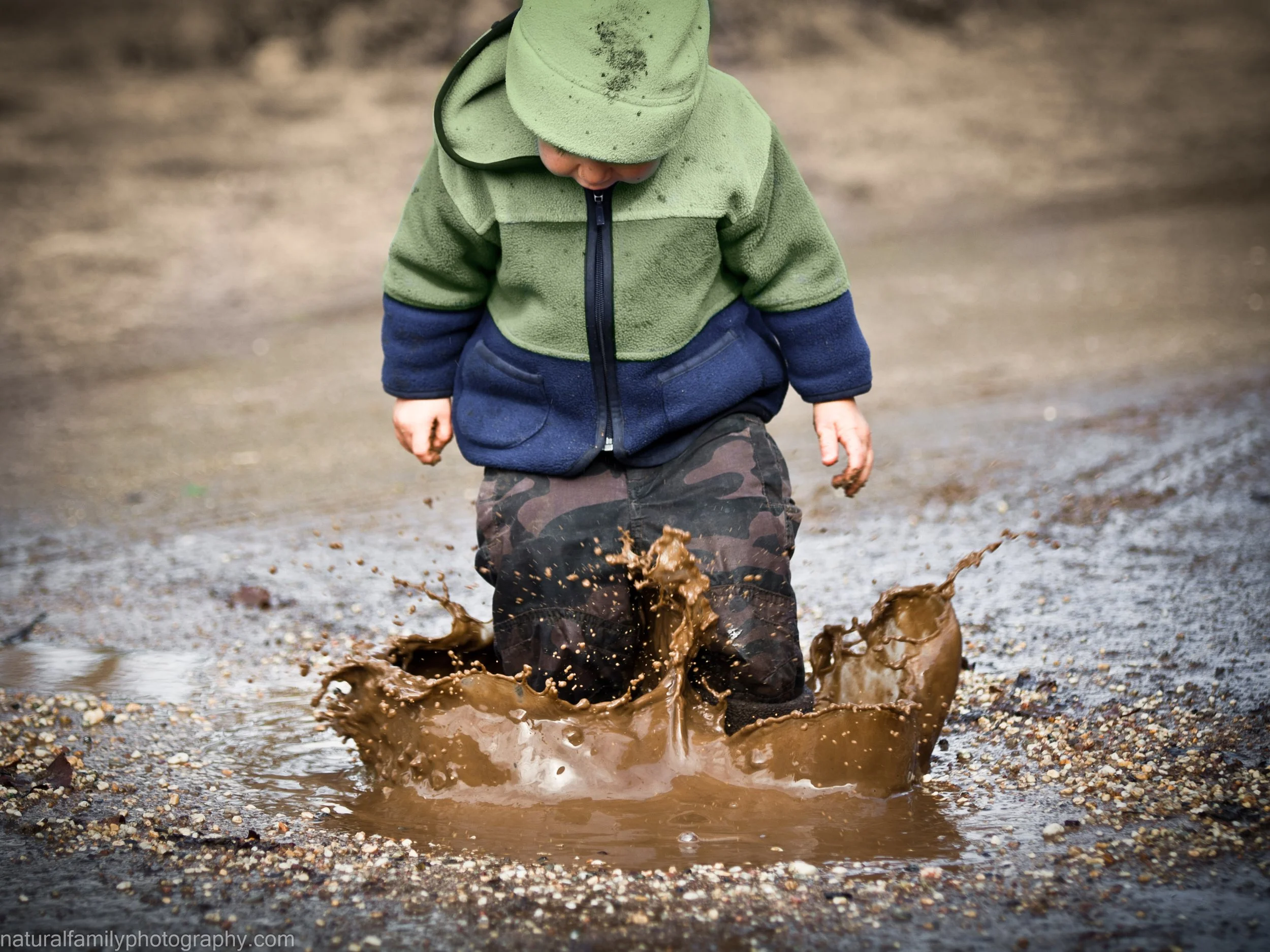 A young boy jumping in a muddy puddle, splashing mud everywhere, wearing a green and navy fleece jacket, camouflage pants, and a green cap. Playful children's portraits by Natural Family Photography, Melbourne.