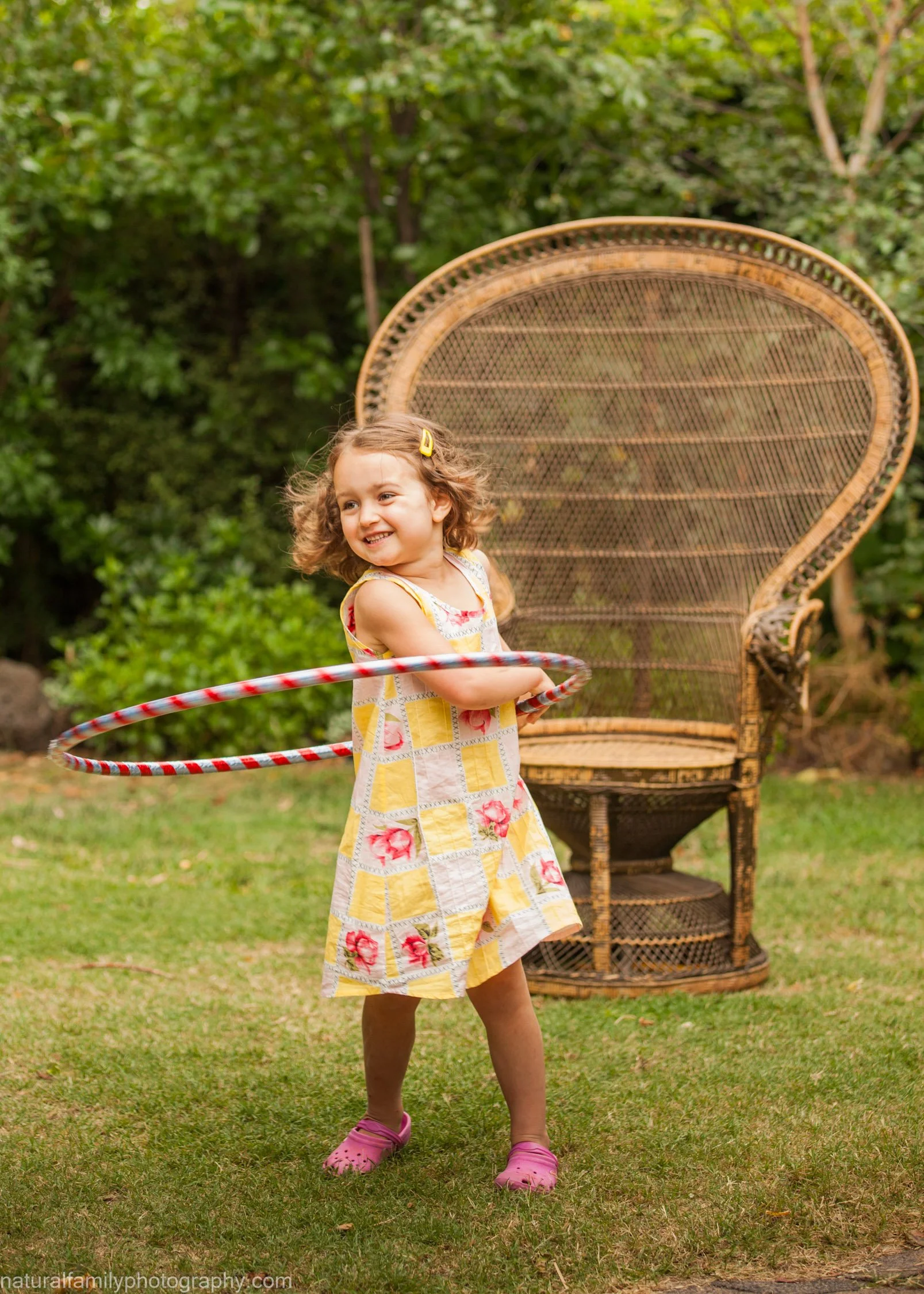 Young girl playing with a hula hoop outdoors in a garden, standing in front of a large wicker chair, wearing a colourful dress and pink shoes, smiling. Playful portrait by Natural Family Photography, Melbourne.
