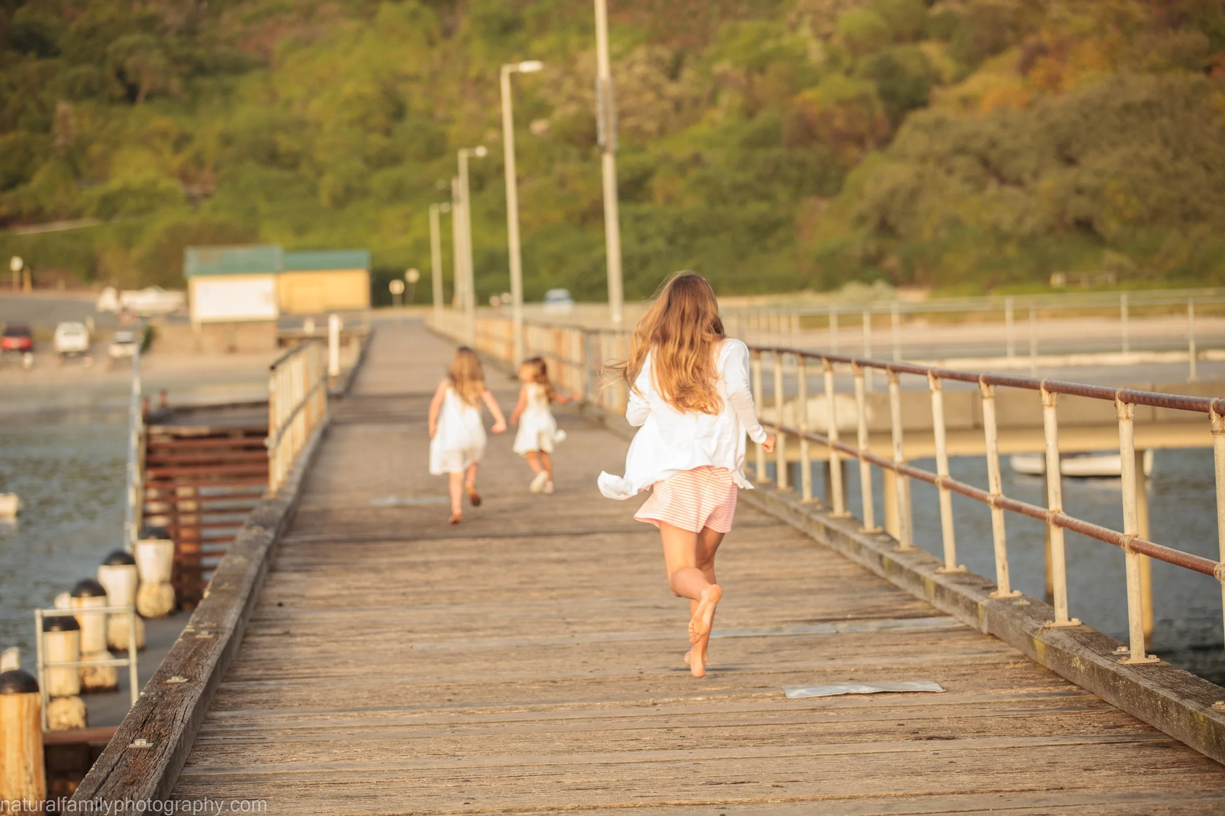 Three girls running on a wooden pier over water with greenery and hills in the background during sunset.