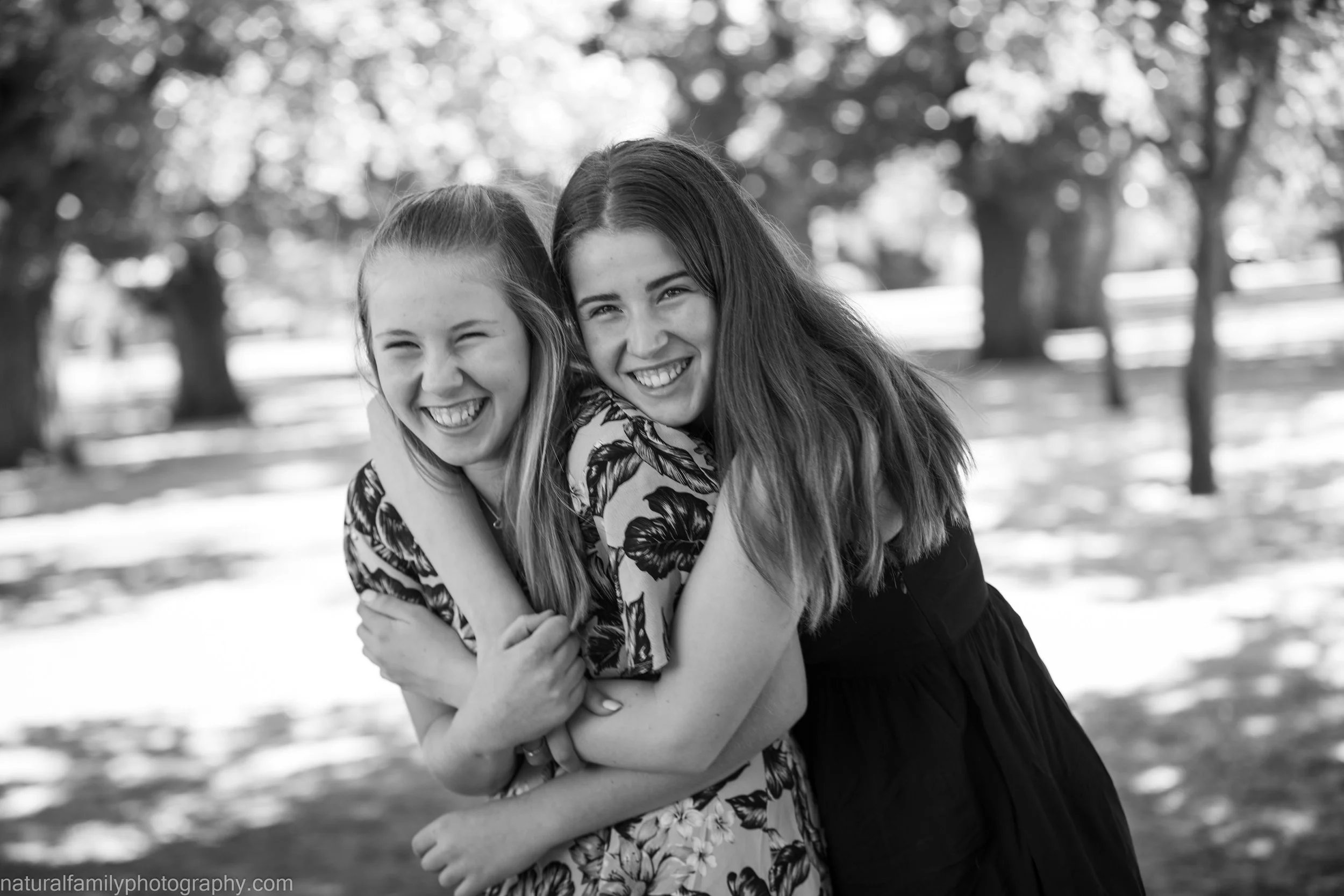 Two smiling girls hugging outdoors in a park, with trees in the background, in black and white.