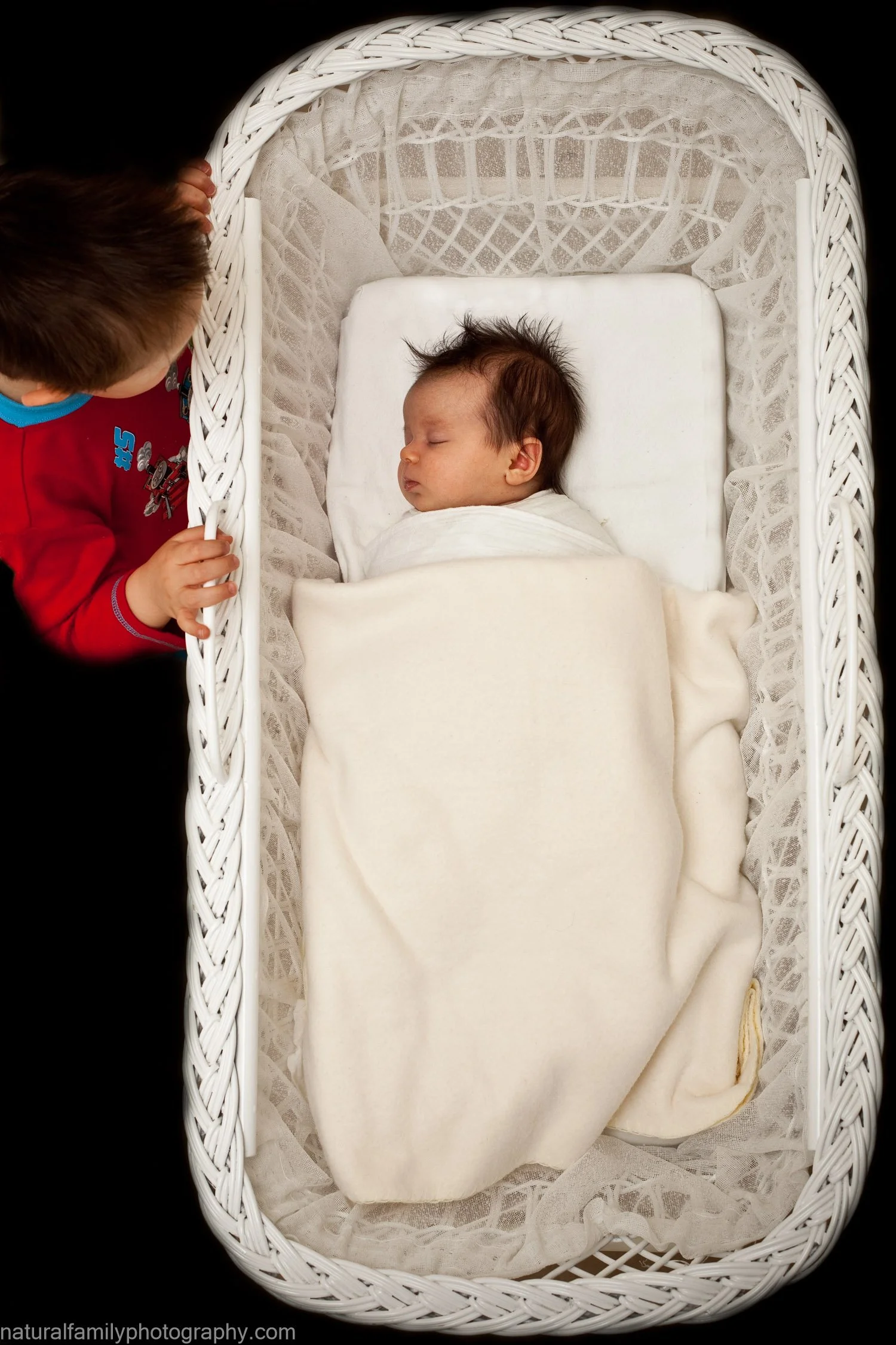 A baby sleeping in a white bassinet with a blanket, viewed from above. A young child reaches in from the side, holding the edge of the bassinet. In-home creative portraiture by Natural Family Photography, Melbourne.