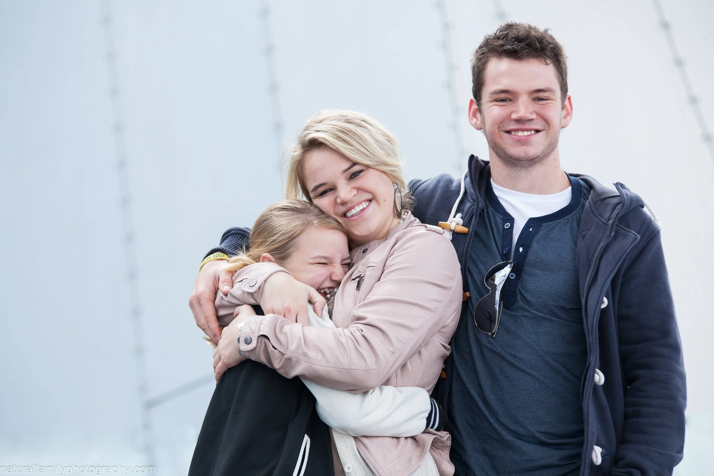 Three people, two women and one man, happily hugging outdoors against a gray sky background.