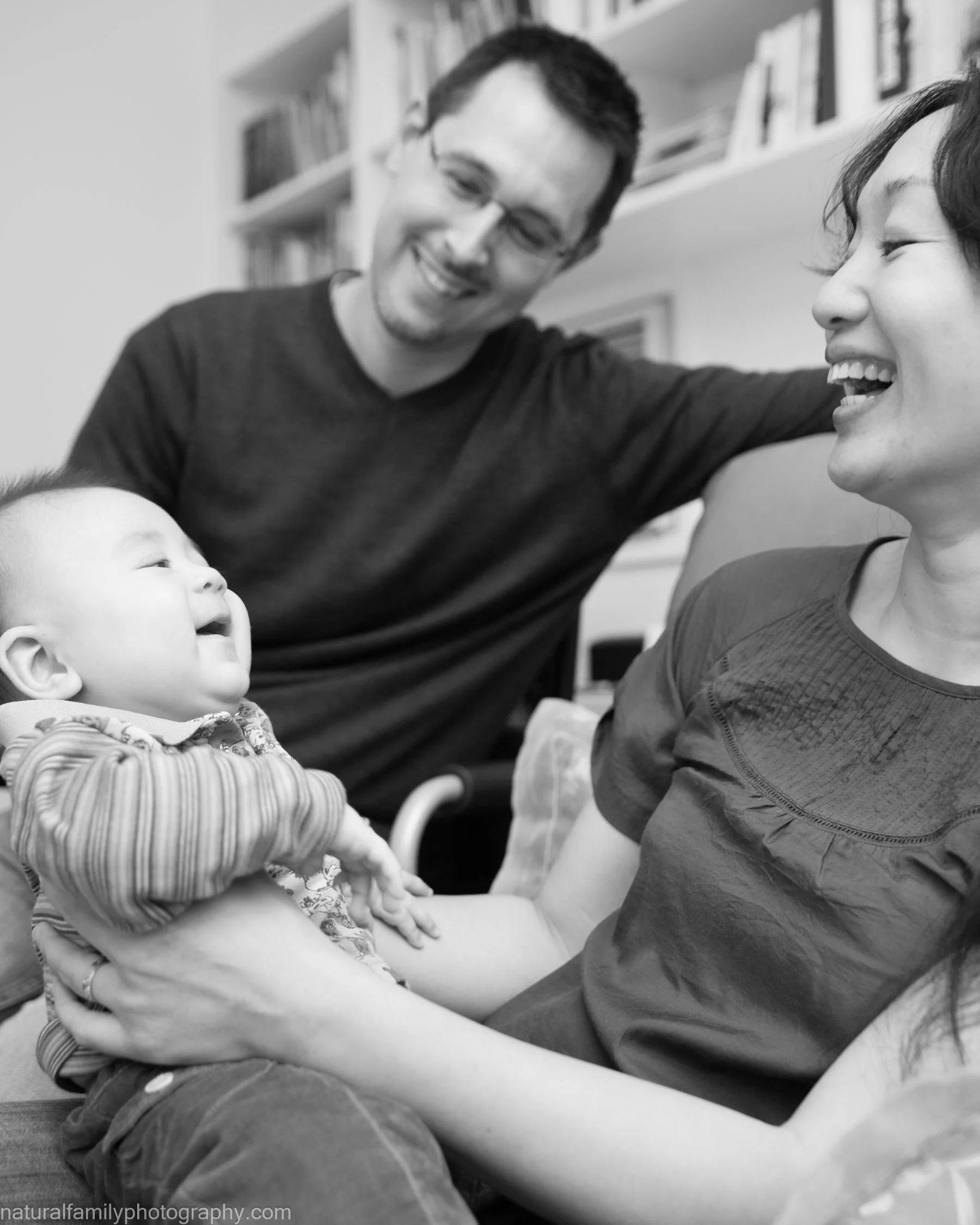 A black and white photo of a family with a mother, father, and baby. The mother is sitting and holding the baby, smiling at her. The father is standing behind them, leaning on a couch, and smiling at the mother and baby. The baby is looking up at the