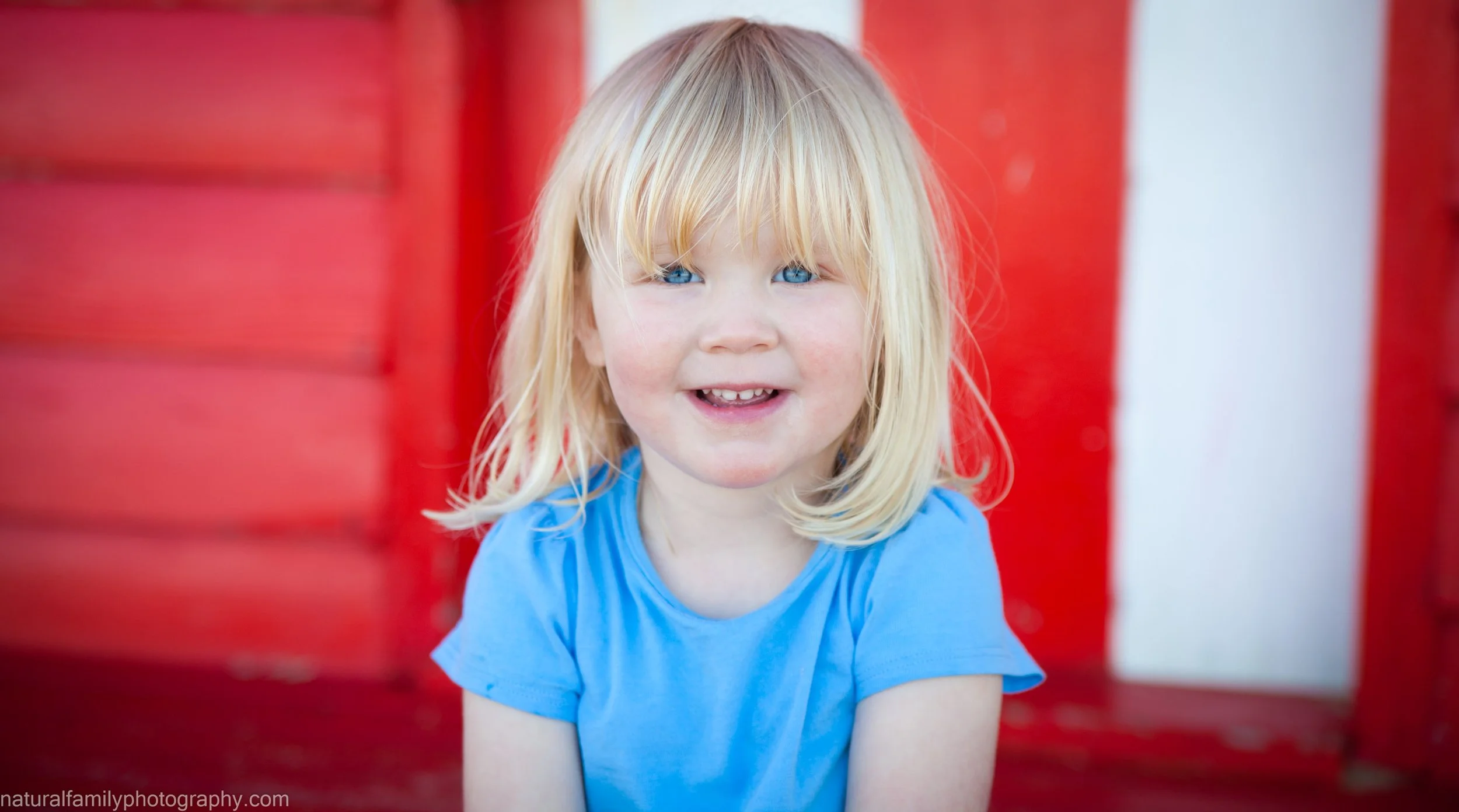 A young girl with blonde hair and blue eyes, smiling at the camera, wearing a blue shirt, standing in front of a red and white background.