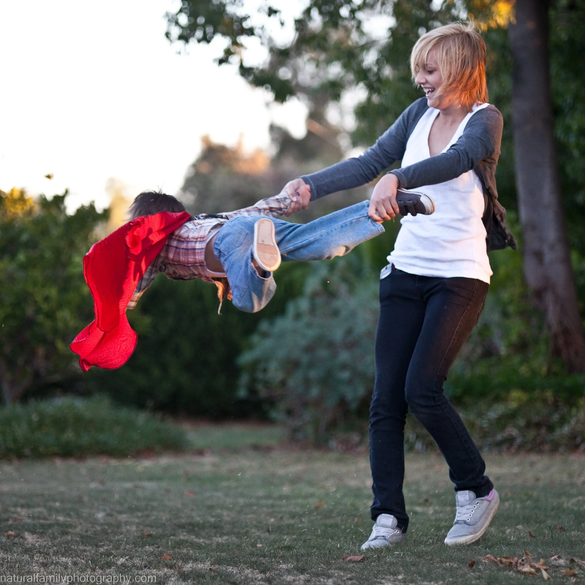 A woman is playing with a young boy outdoors, swinging him around by his arm, both smiling and enjoying themselves. Child wears a cape. Playful portrait by Natural Family Photography, Melbourne.