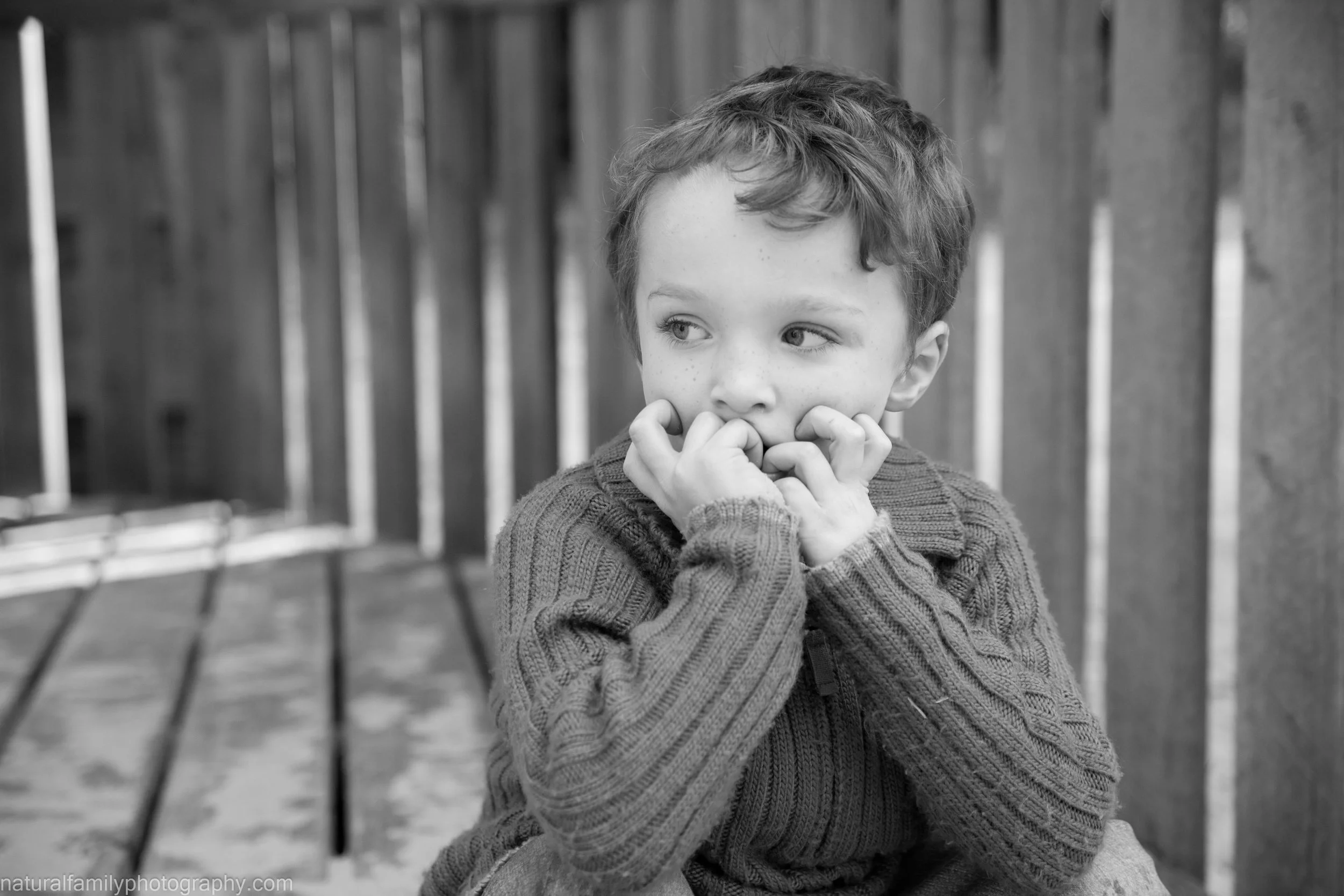 A young boy with curly hair and freckles sits on a bench near a wooden fence, biting his nails and looking to the side with a thoughtful expression.