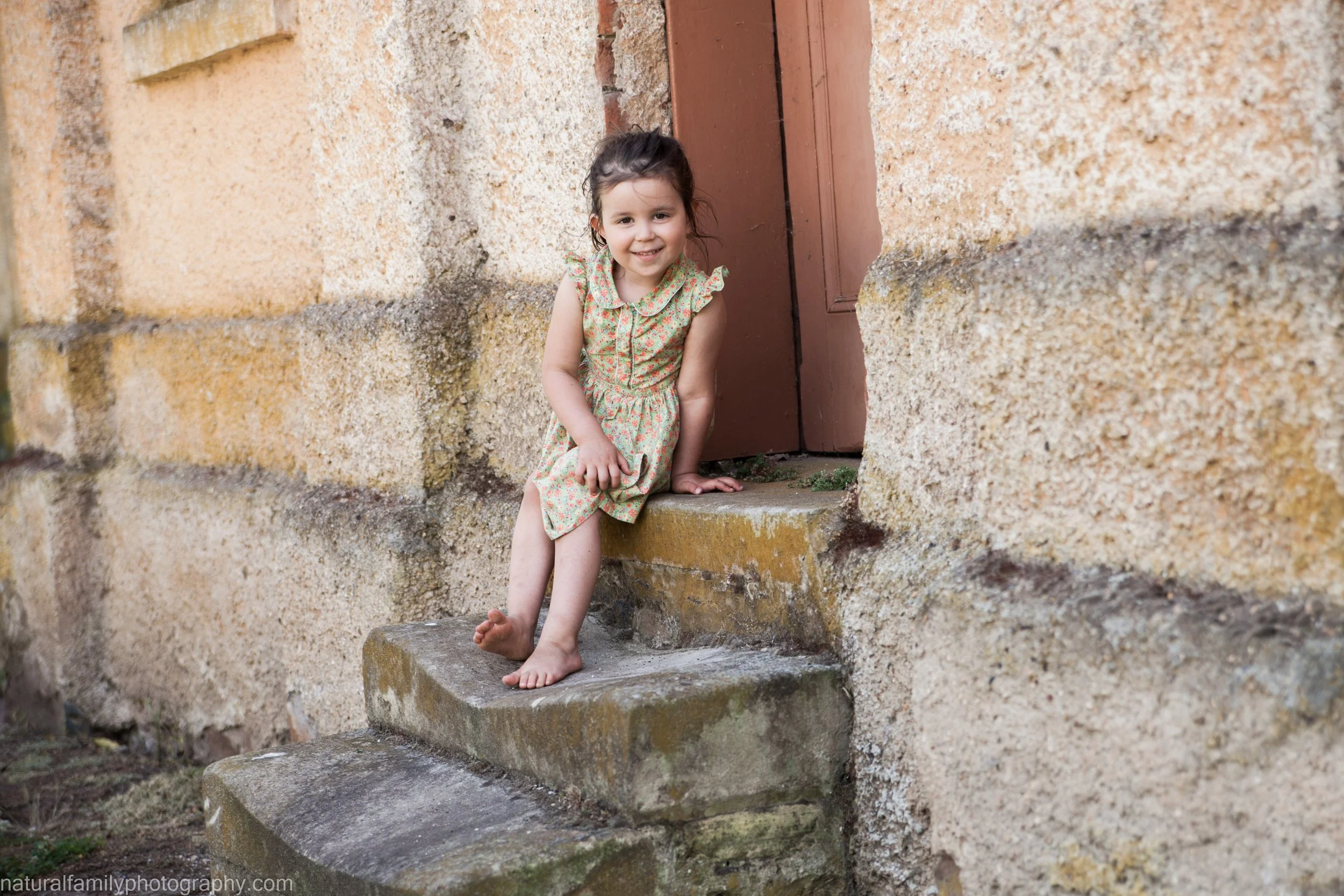 A young girl with dark hair in a braid, wearing a floral dress, smiling, sitting barefoot on stone steps outside a rustic building with a textured yellowish wall and brown wooden door.