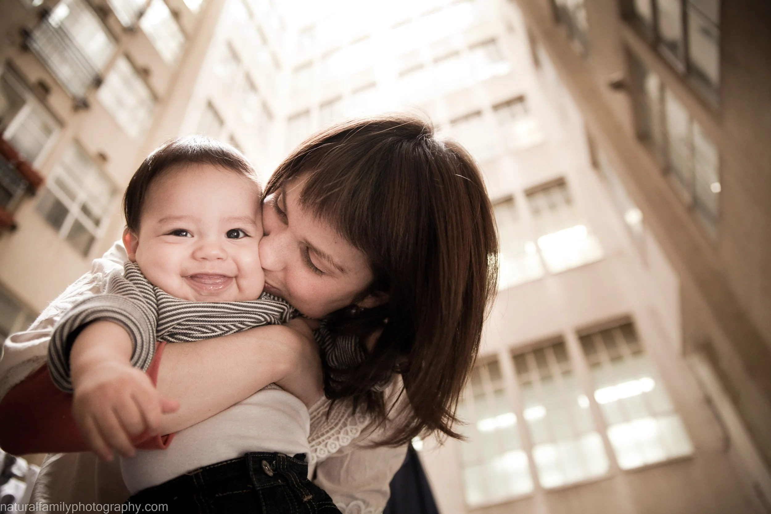 A woman holding a smiling baby in an indoor setting with tall windows and apartment buildings in the background.
